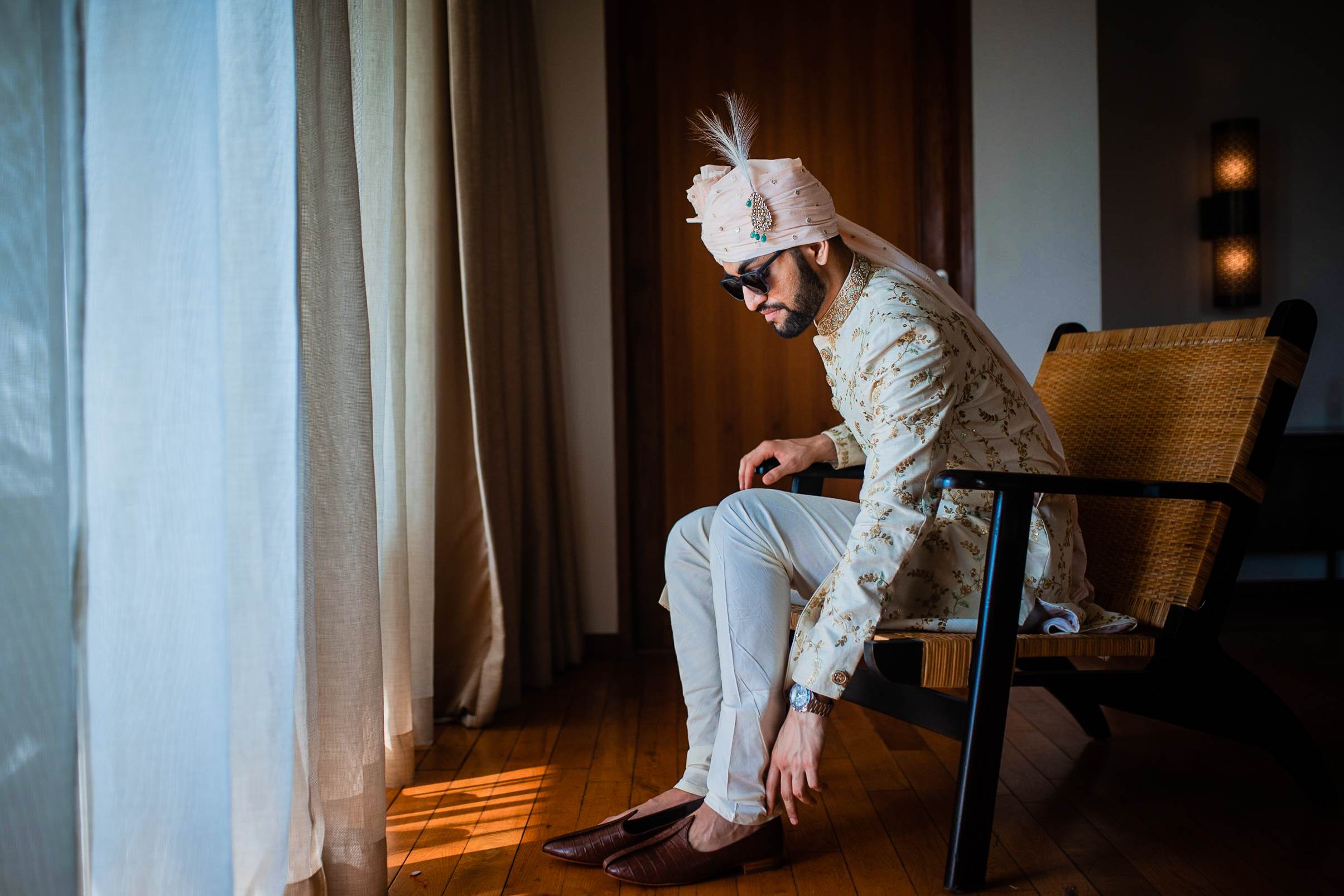 Srikant seated in a woven chair, looking down at his phone, wearing patterned shirt and white headwrap in warm indoor light