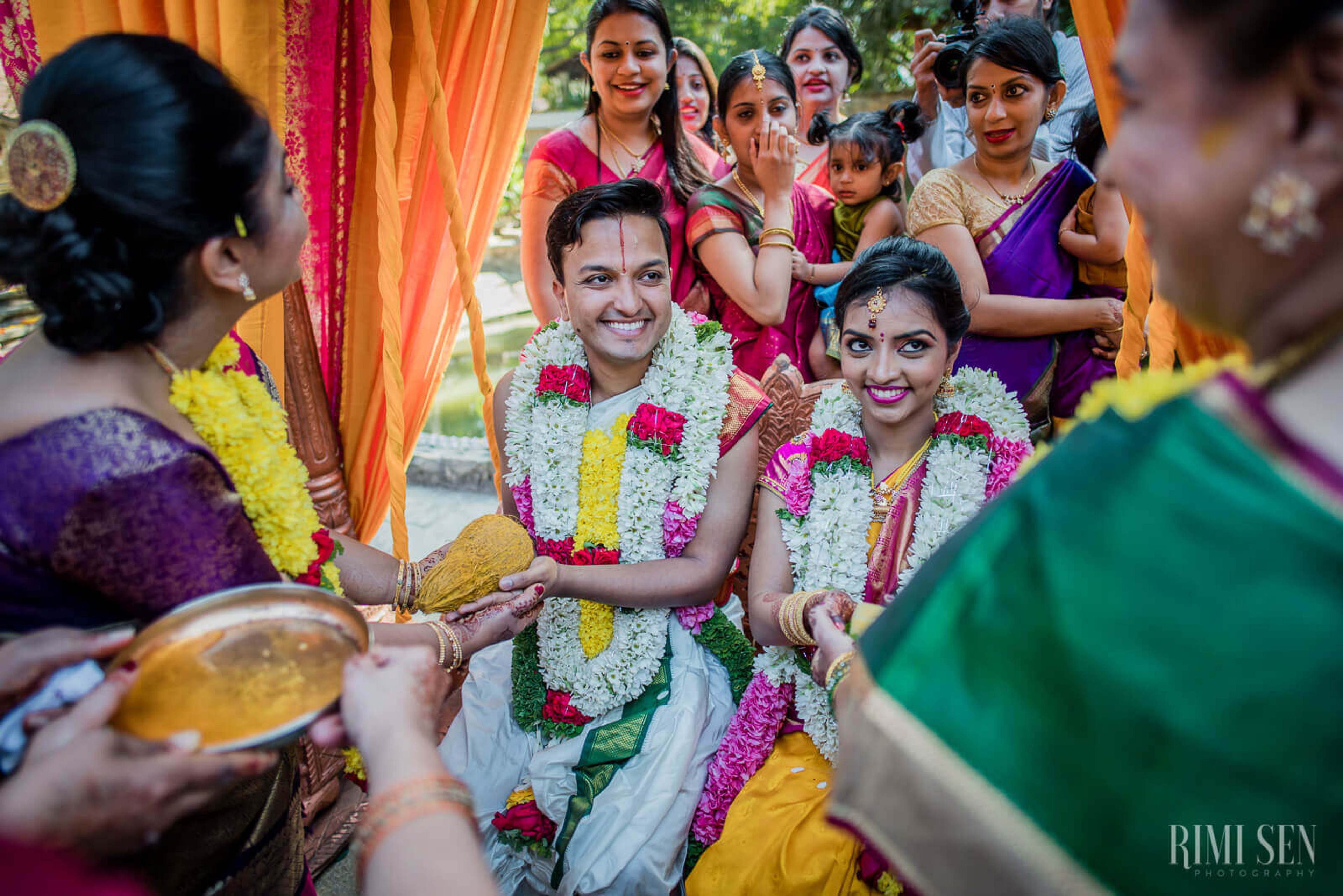 Pooja and Aniket wear pink and white floral garlands, smiling amid colorful fabrics and family at their Bangalore wedding ceremony