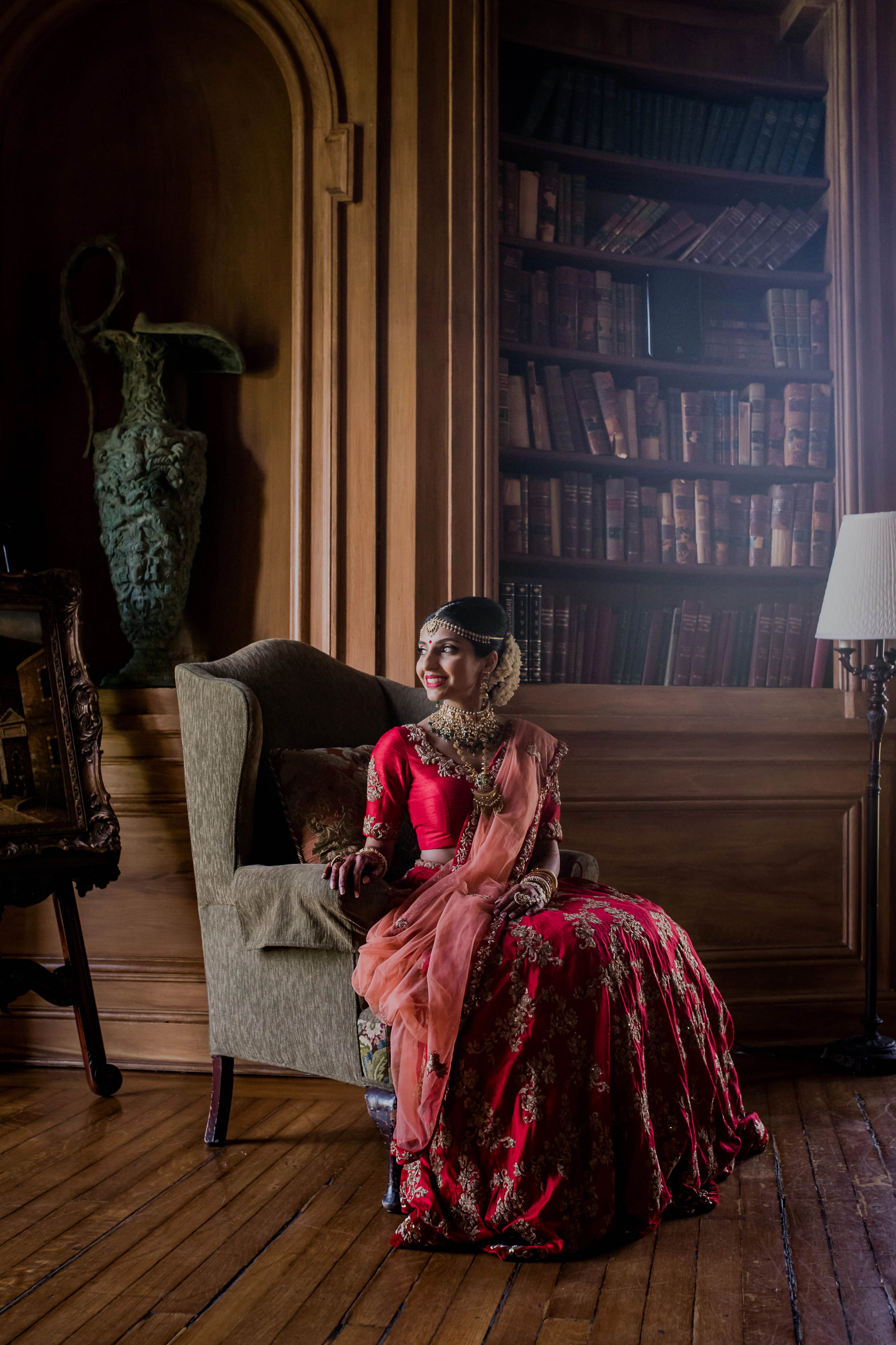 Woman in red and gold embroidered wedding attire seated in cream chair, library backdrop with books and vintage decor