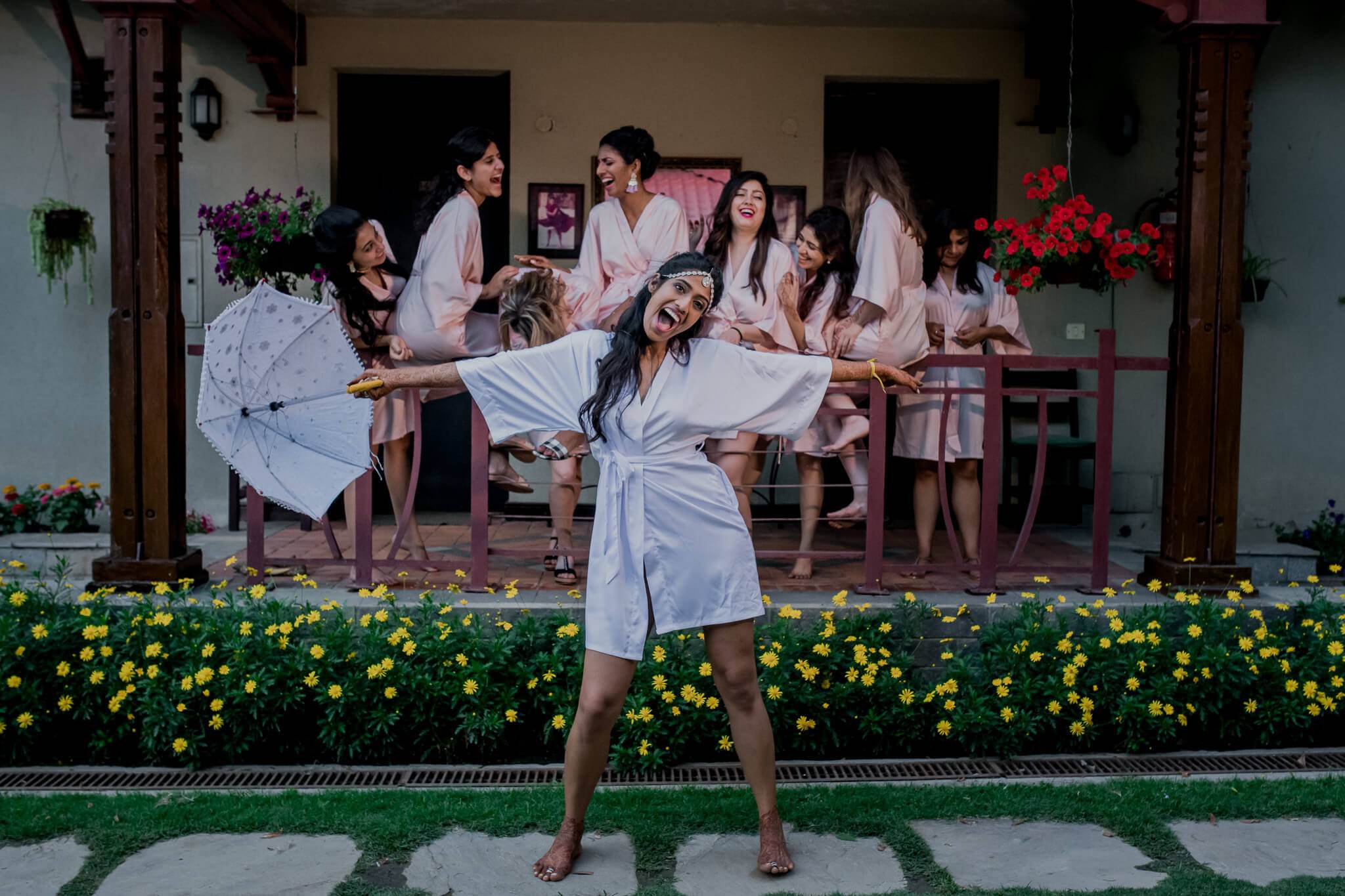 Anu in white robe twirls with parasol before bridesmaids in Nepal courtyard with yellow flowers