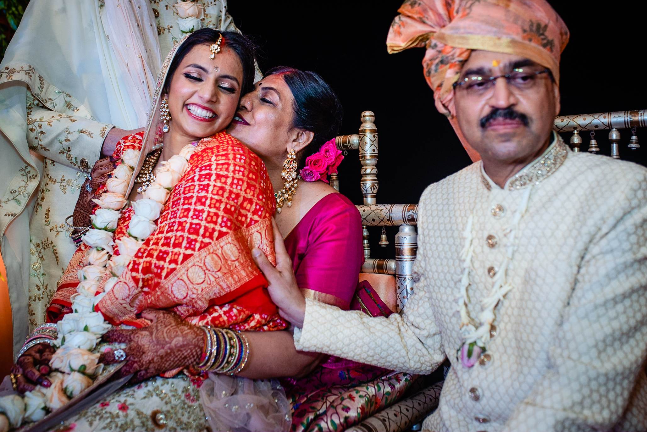 Avani in red wedding lehenga with flower garland, embraced by family member in magenta, Srikant in cream sherwani beside them