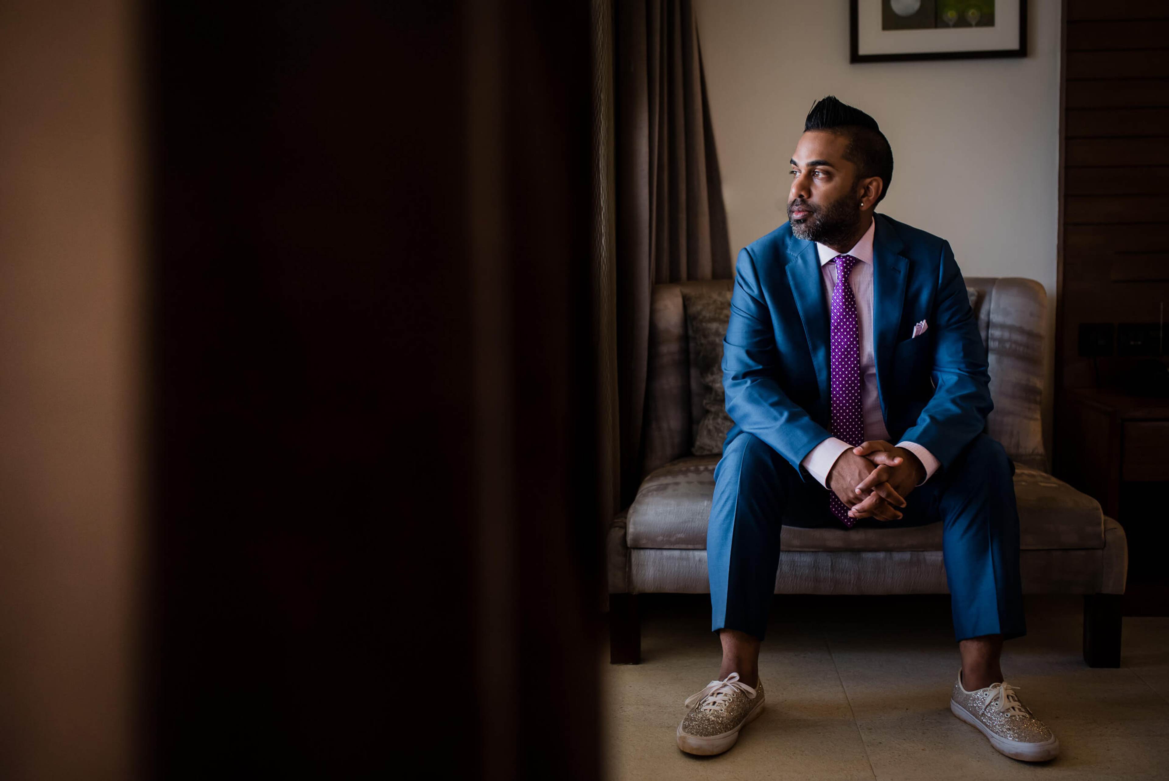 Groom in blue suit and purple tie sitting on chair, looking thoughtful in indoor setting