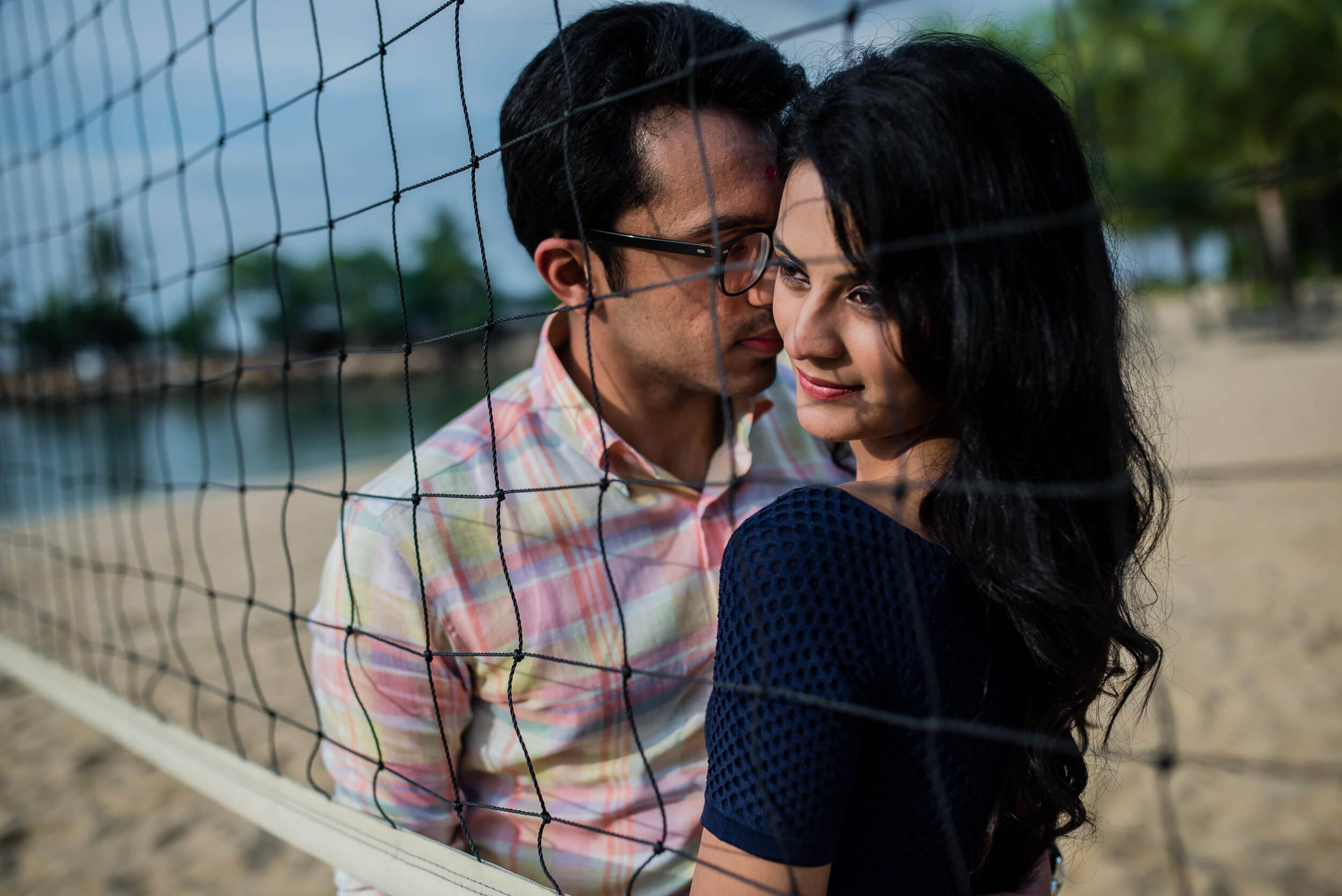 Yash and Disha pose together behind a volleyball net on a sunny beach in Singapore