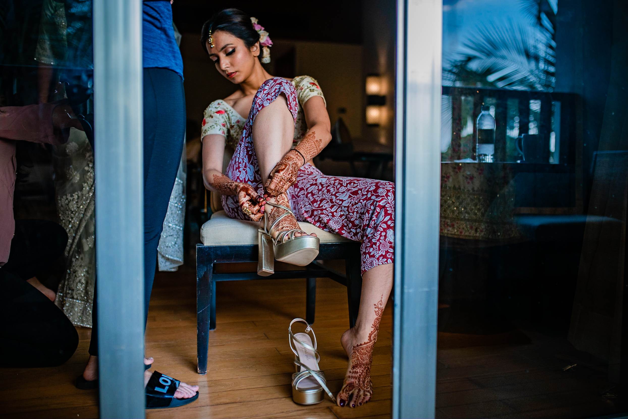 Avani sits on a chair adjusting her heeled sandal, wearing an ornate purple patterned outfit and henna, indoors in Goa