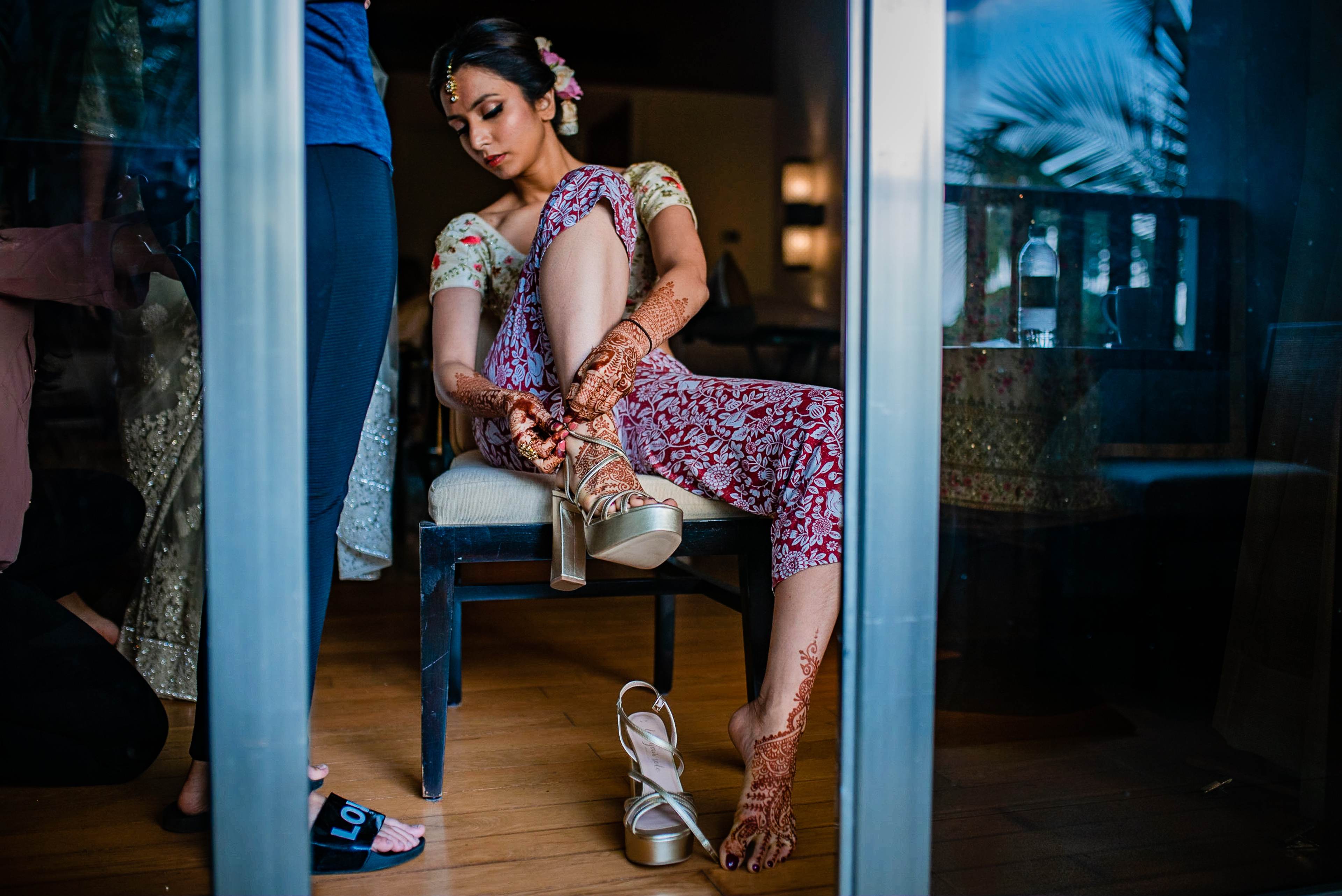 Avani sits on a chair adjusting her heeled sandal, wearing an ornate purple patterned outfit and henna, indoors in Goa