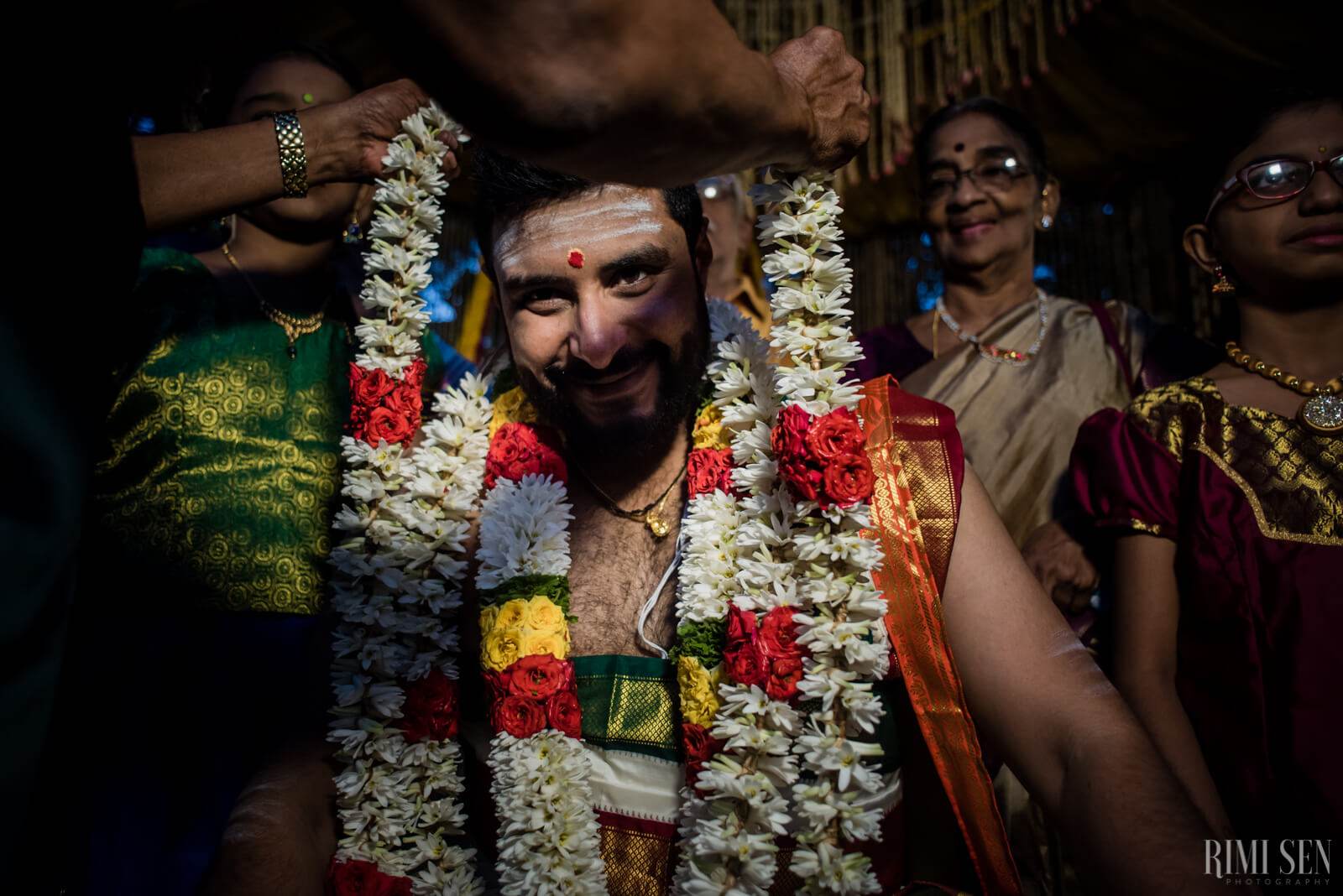 Sridhar wears traditional flower garlands and red tilak, surrounded by smiling family in colorful silk at Bangalore wedding celebration