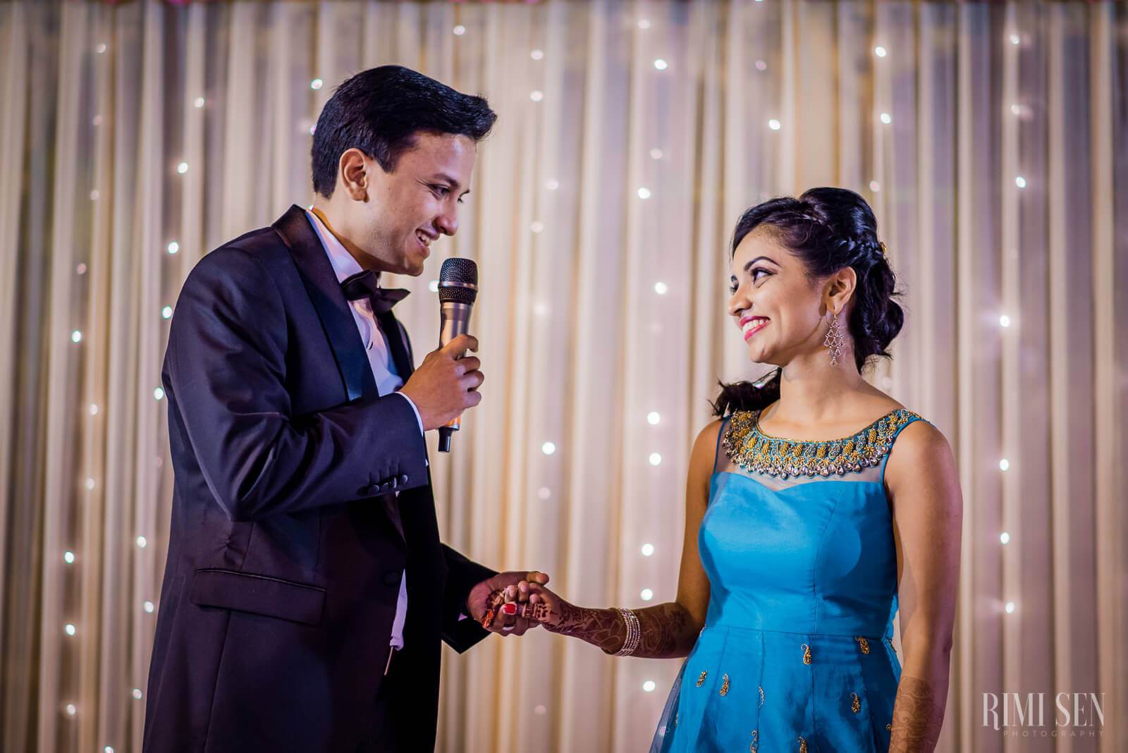 Aniket holds microphone speaking to Pooja in blue dress before twinkle-lit curtain backdrop