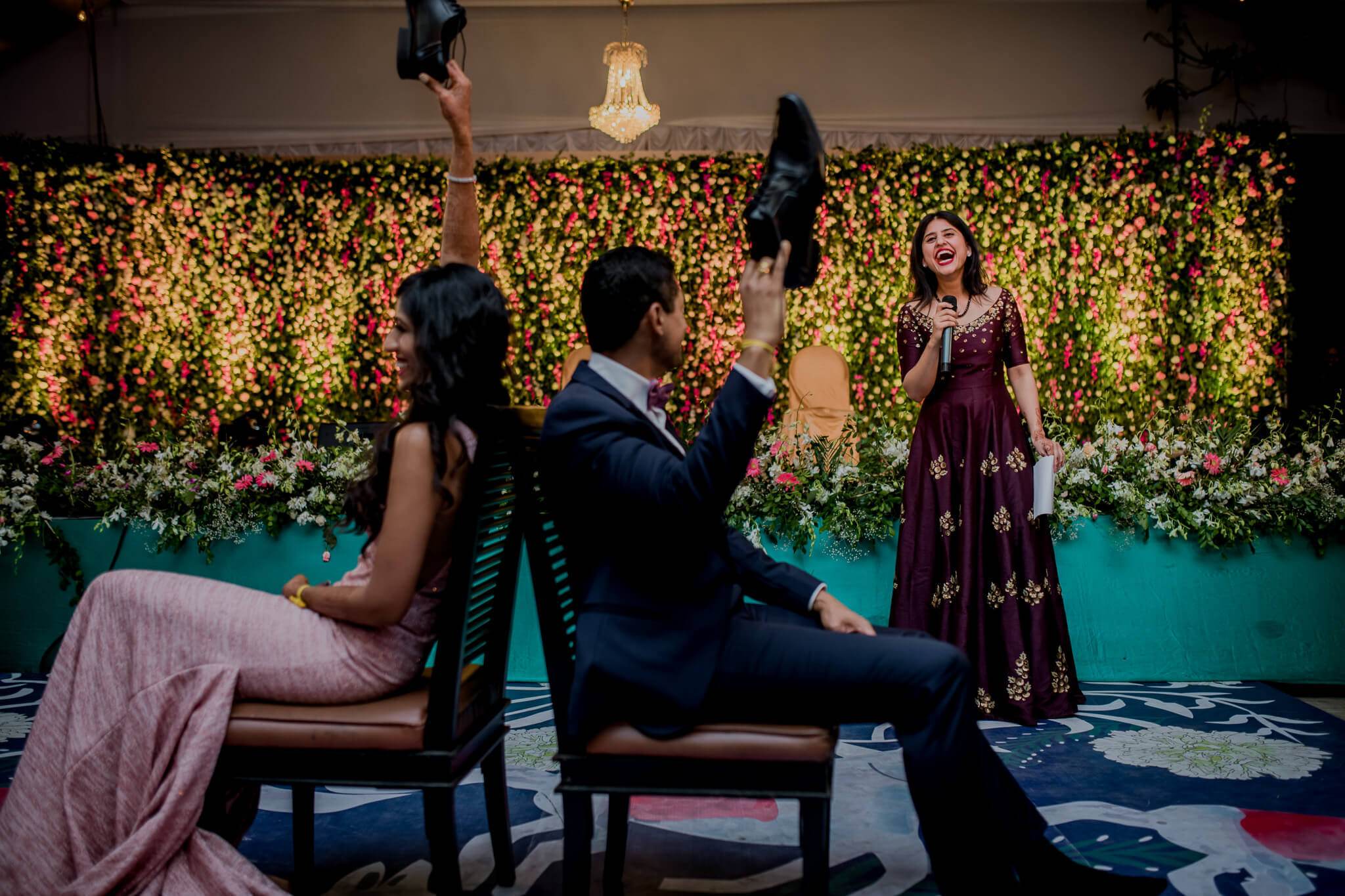 Anu and Raghu sit playfully on chairs before a lush floral backdrop, tossing shoes in celebration