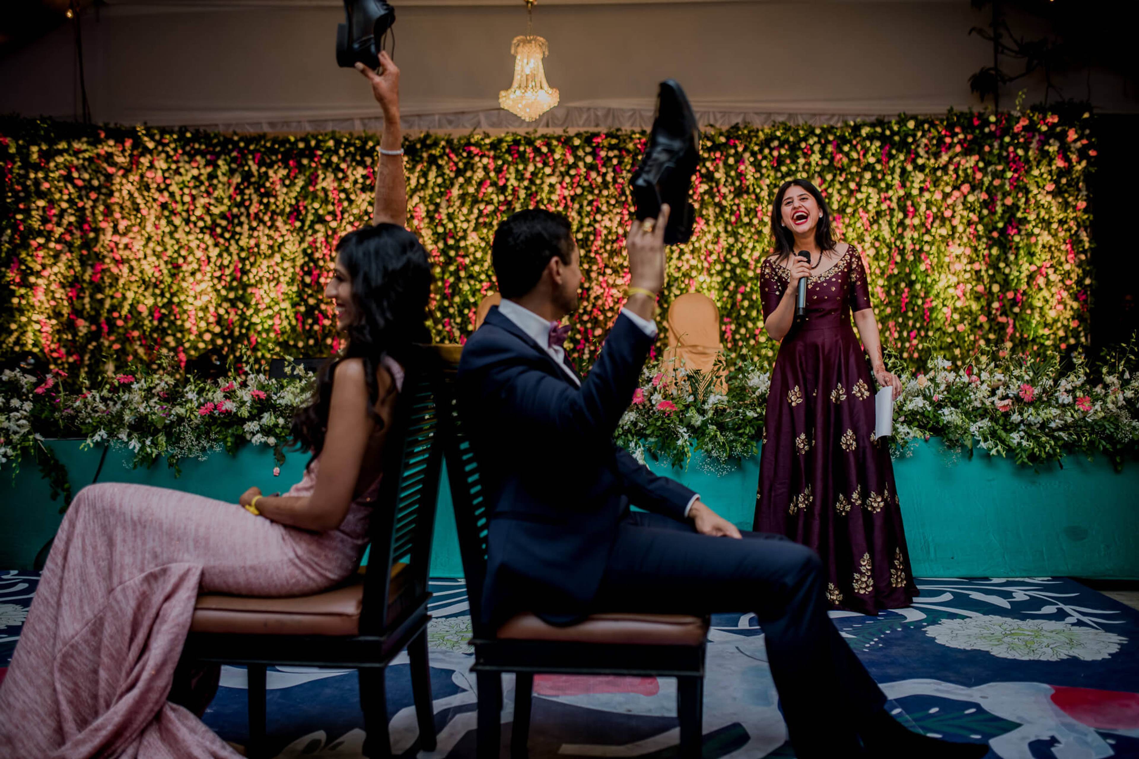 Anu and Raghu sit playfully on chairs before a lush floral backdrop, tossing shoes in celebration