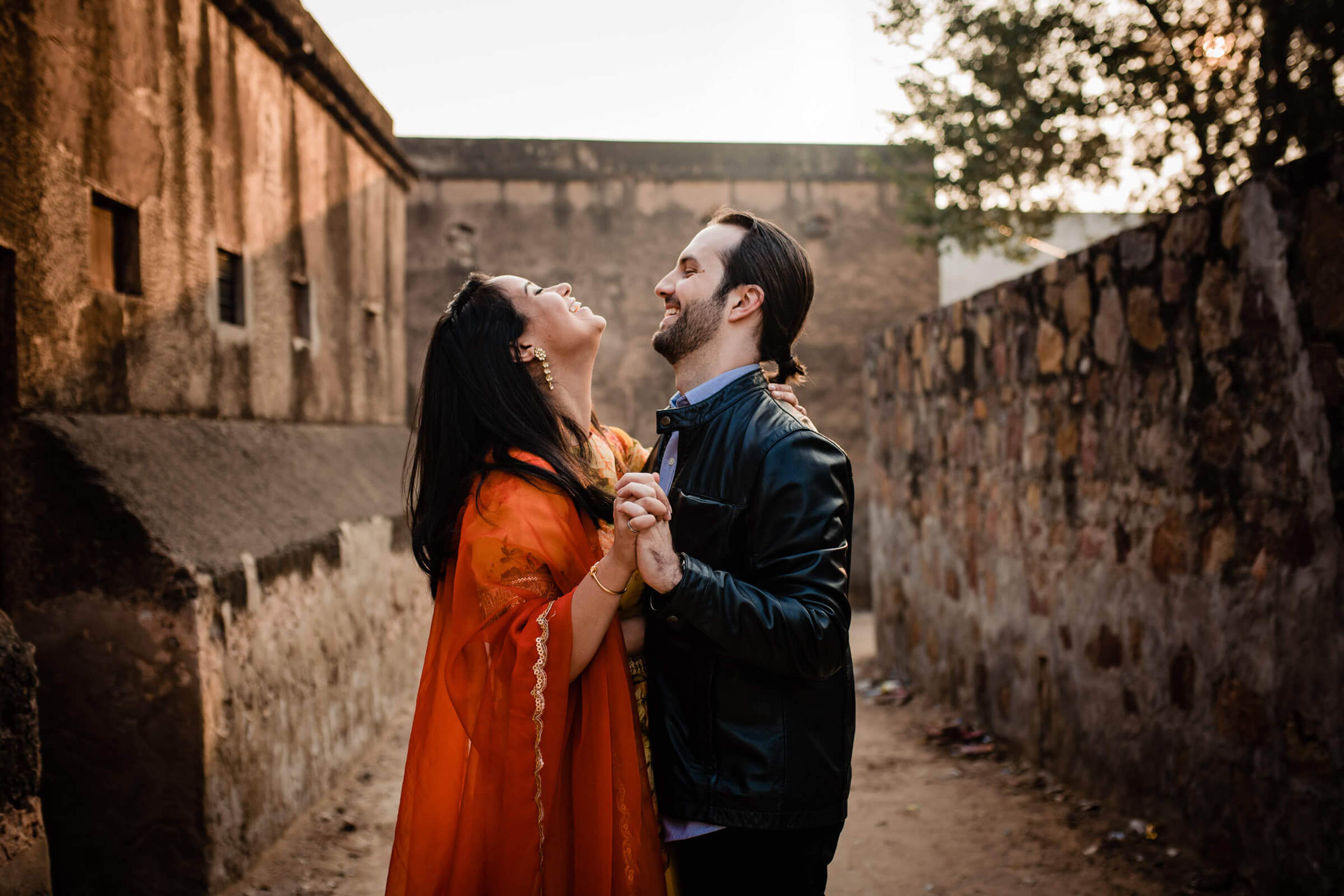 Maitrayee in vibrant orange dress and Eric in navy jacket smile in narrow Rajasthan alleyway with stone walls