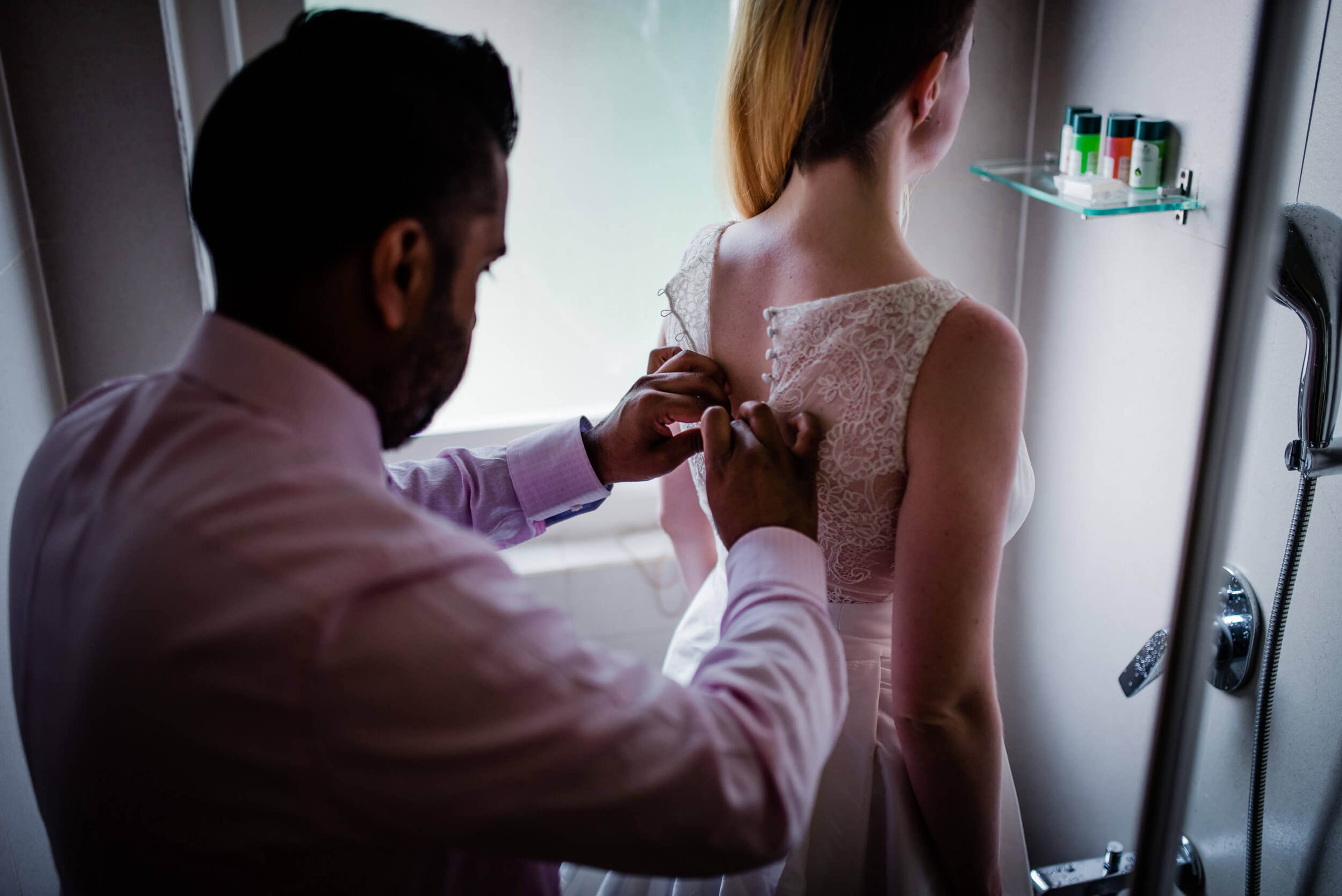 Man in pink shirt fastening Sarah's lace dress back in Goa bathroom, natural window light