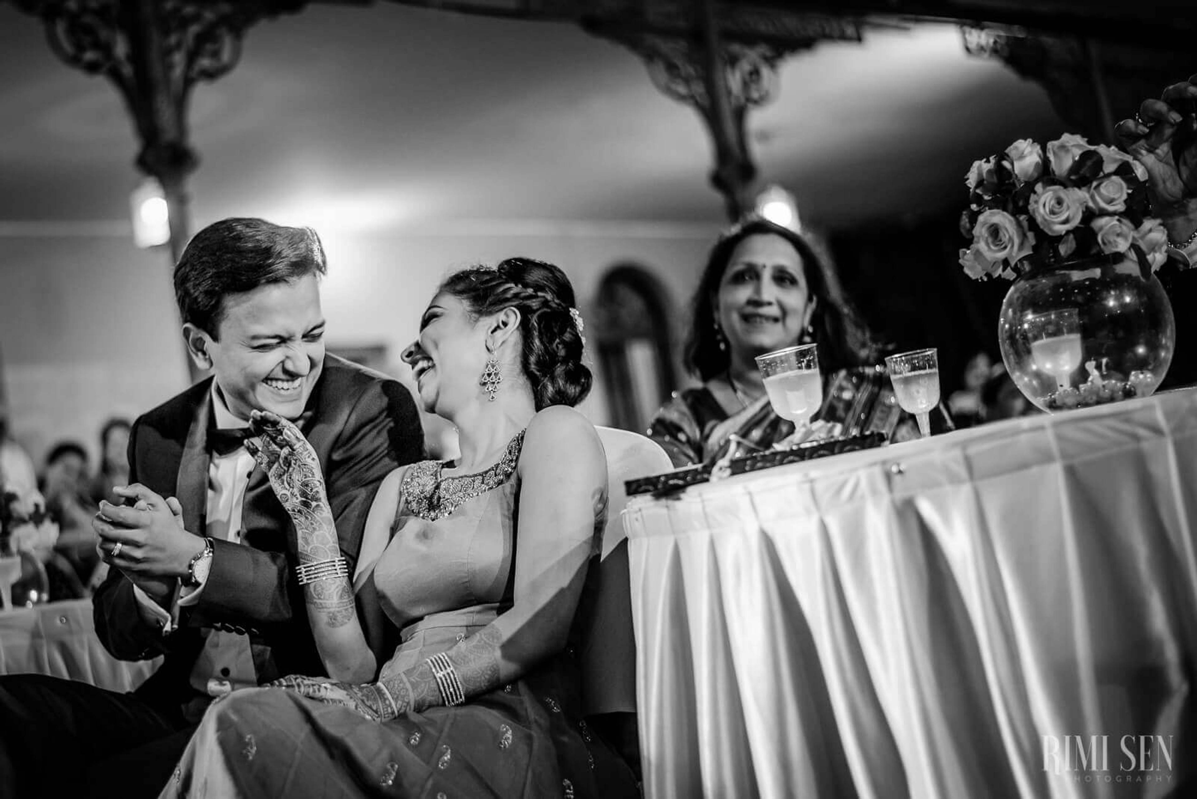 Pooja and Aniket laugh together at their Bangalore wedding reception, seated at the head table with guests and floral arrangements nearby