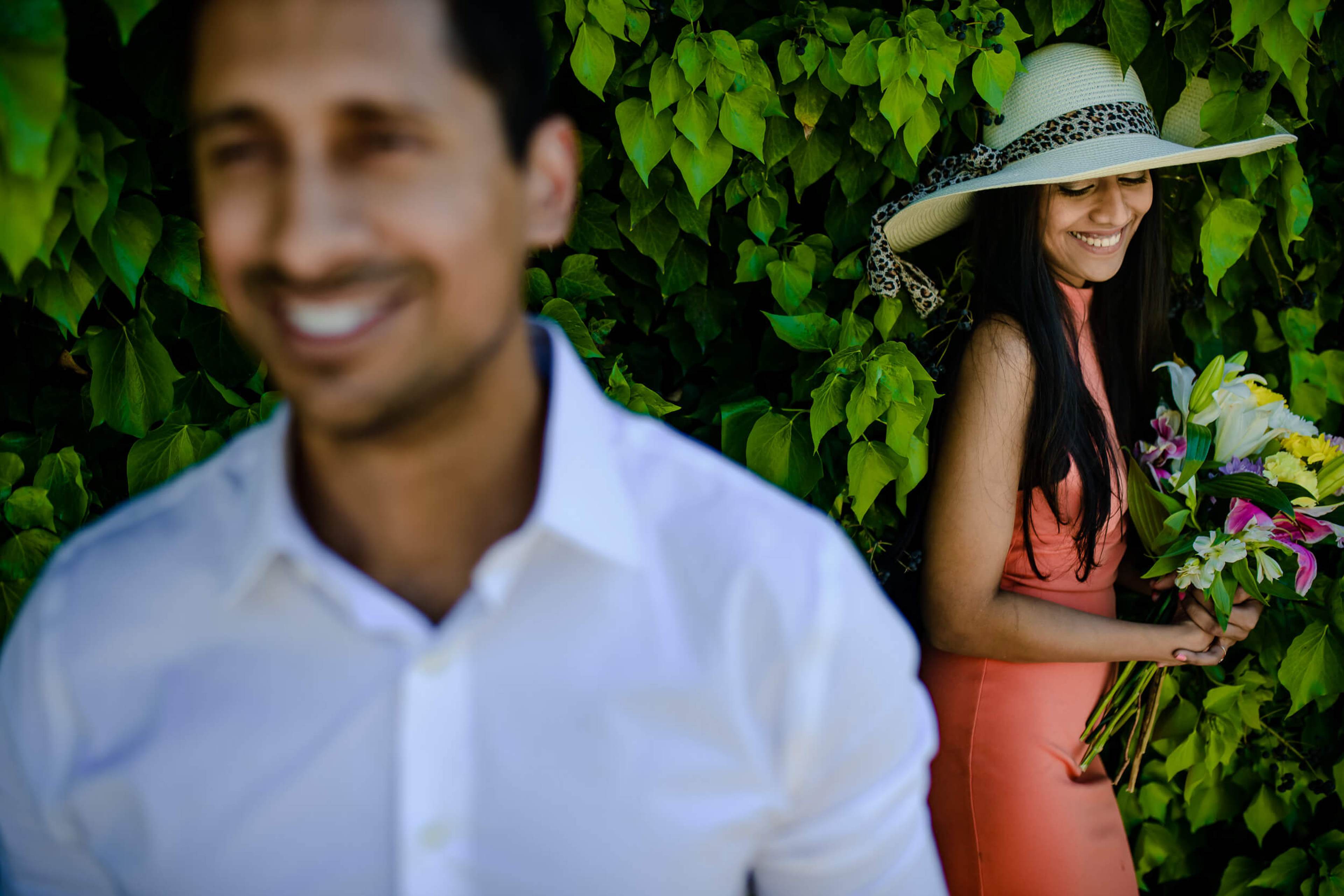 Anu and Raghu smile among lush green foliage, she in coral dress and wide-brimmed hat, he in white shirt
