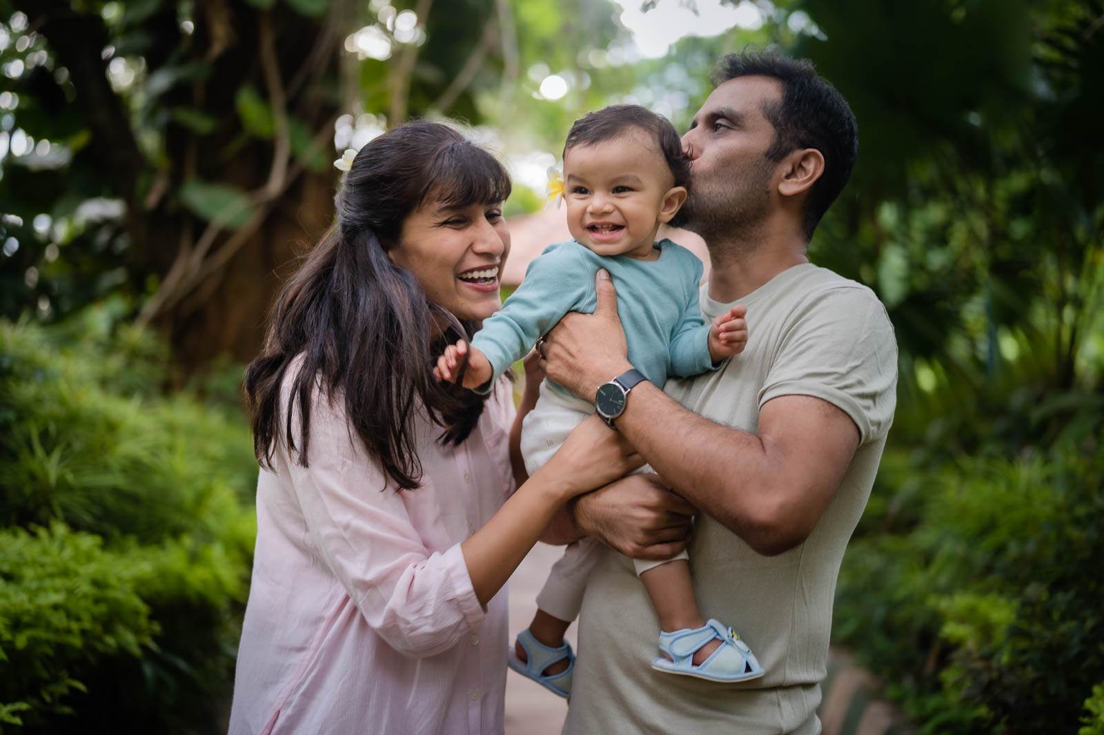 Parents holding smiling toddler among lush green foliage, bright natural light filtering through trees