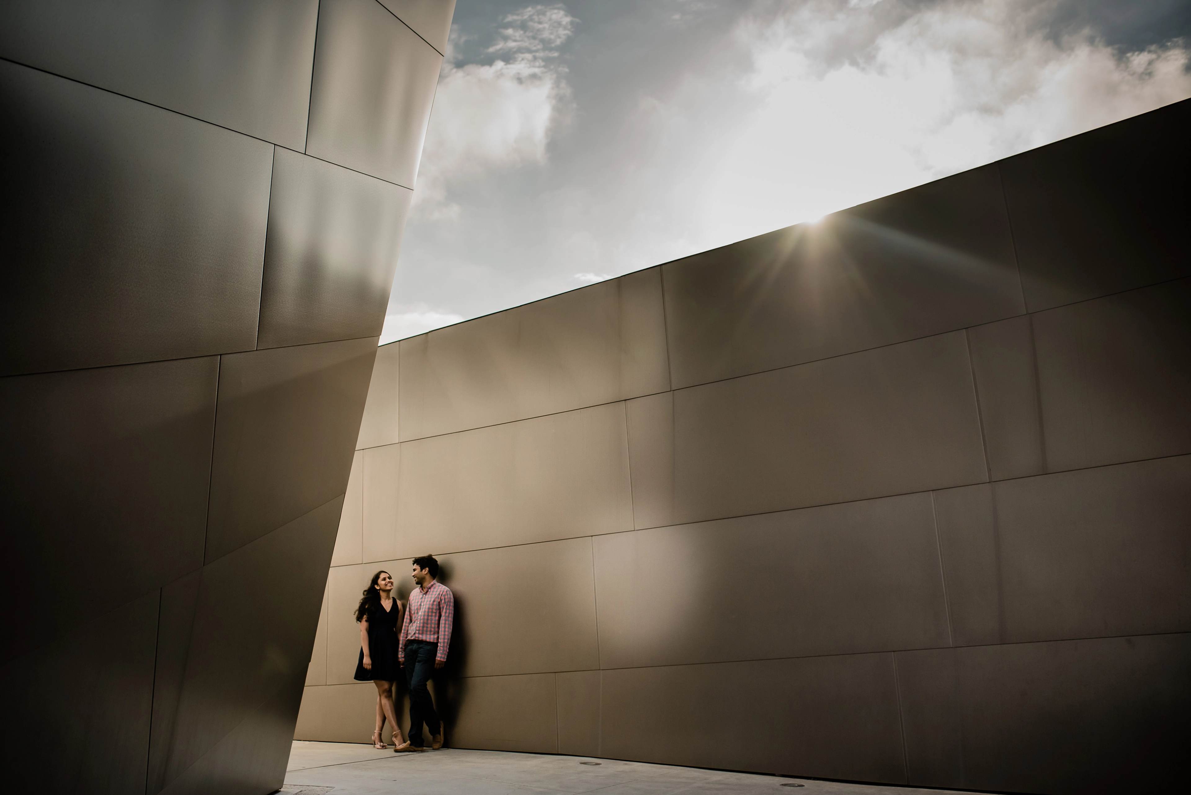 Two people stand against a modern concrete wall under bright sunlight and cloudy sky