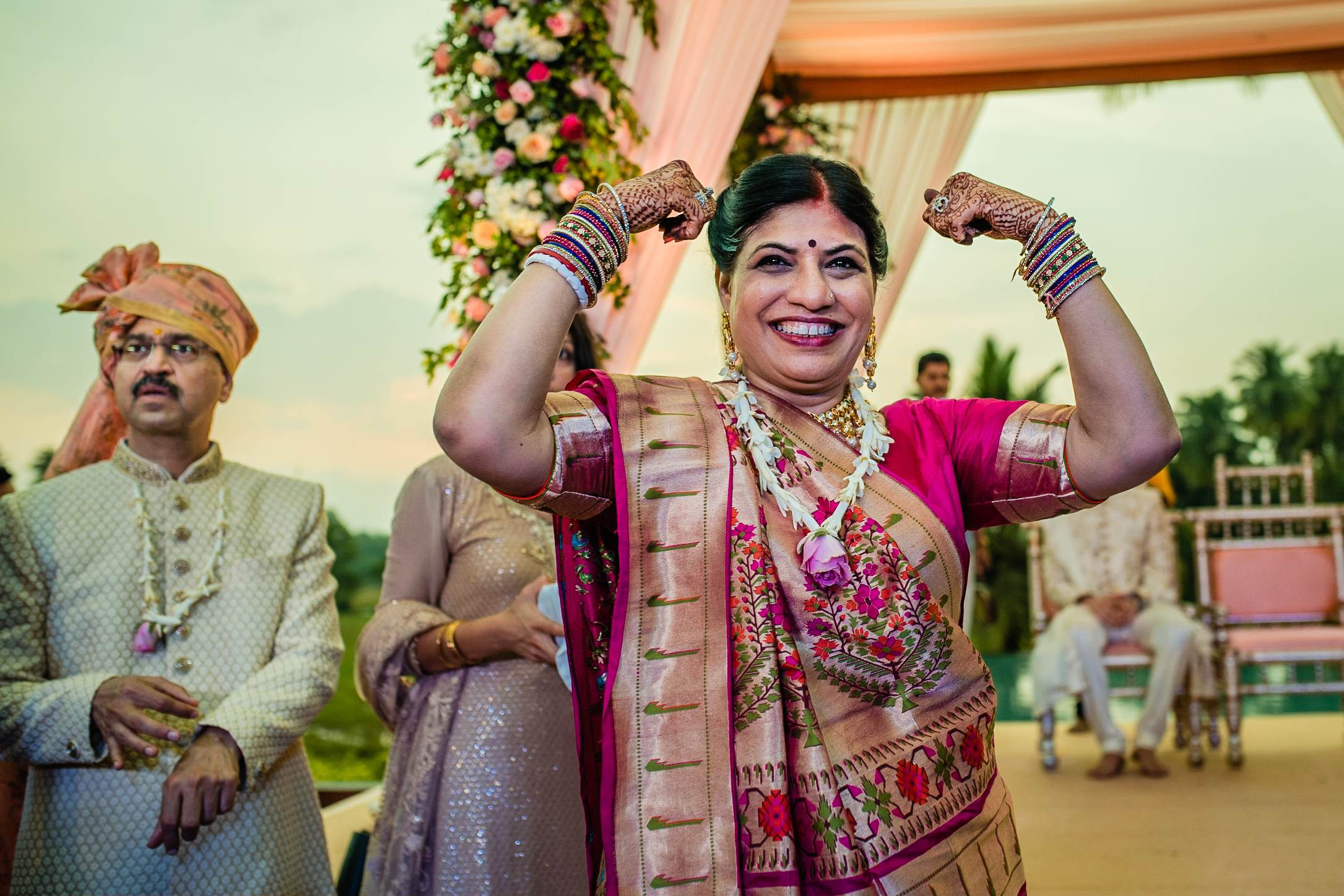 Avani celebrates in traditional Indian wedding attire with raised arms, Srikant beside her, floral arch and decorated venue in Goa