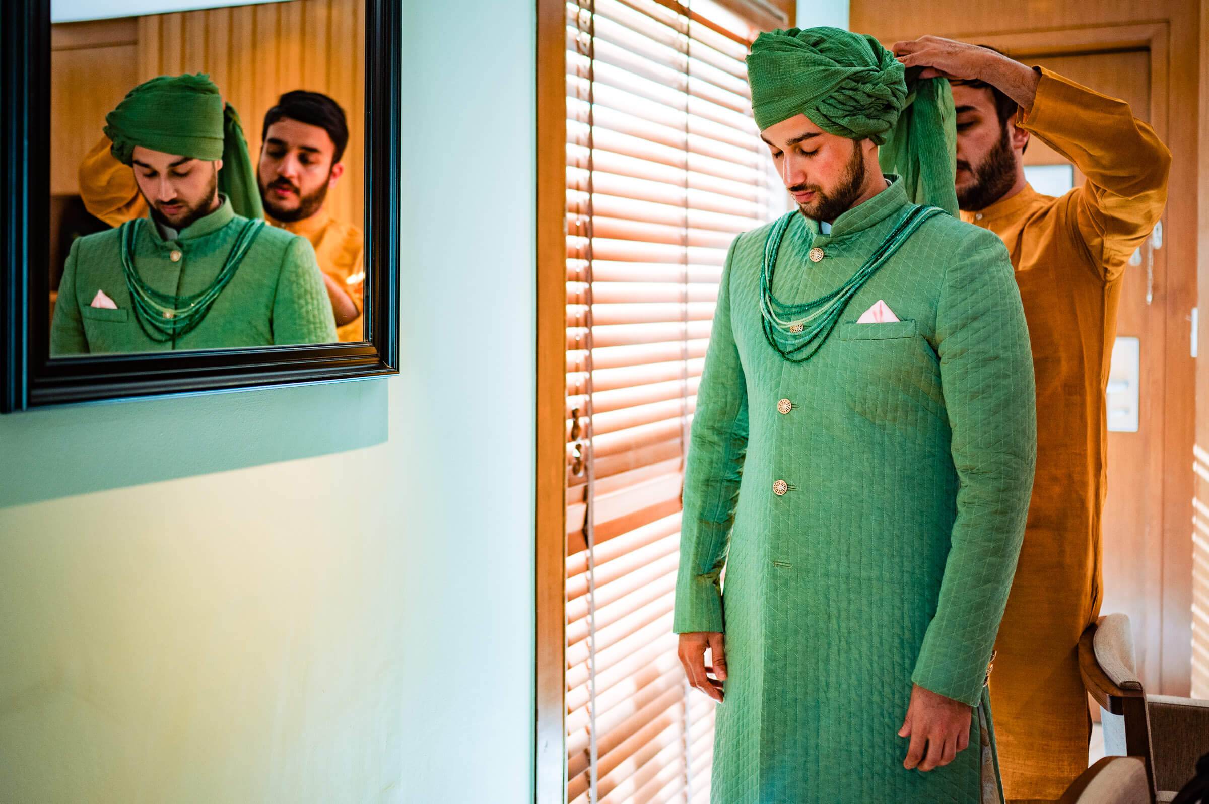 Shreya and Yash in green traditional attire with draped headwear, standing indoors by window blinds