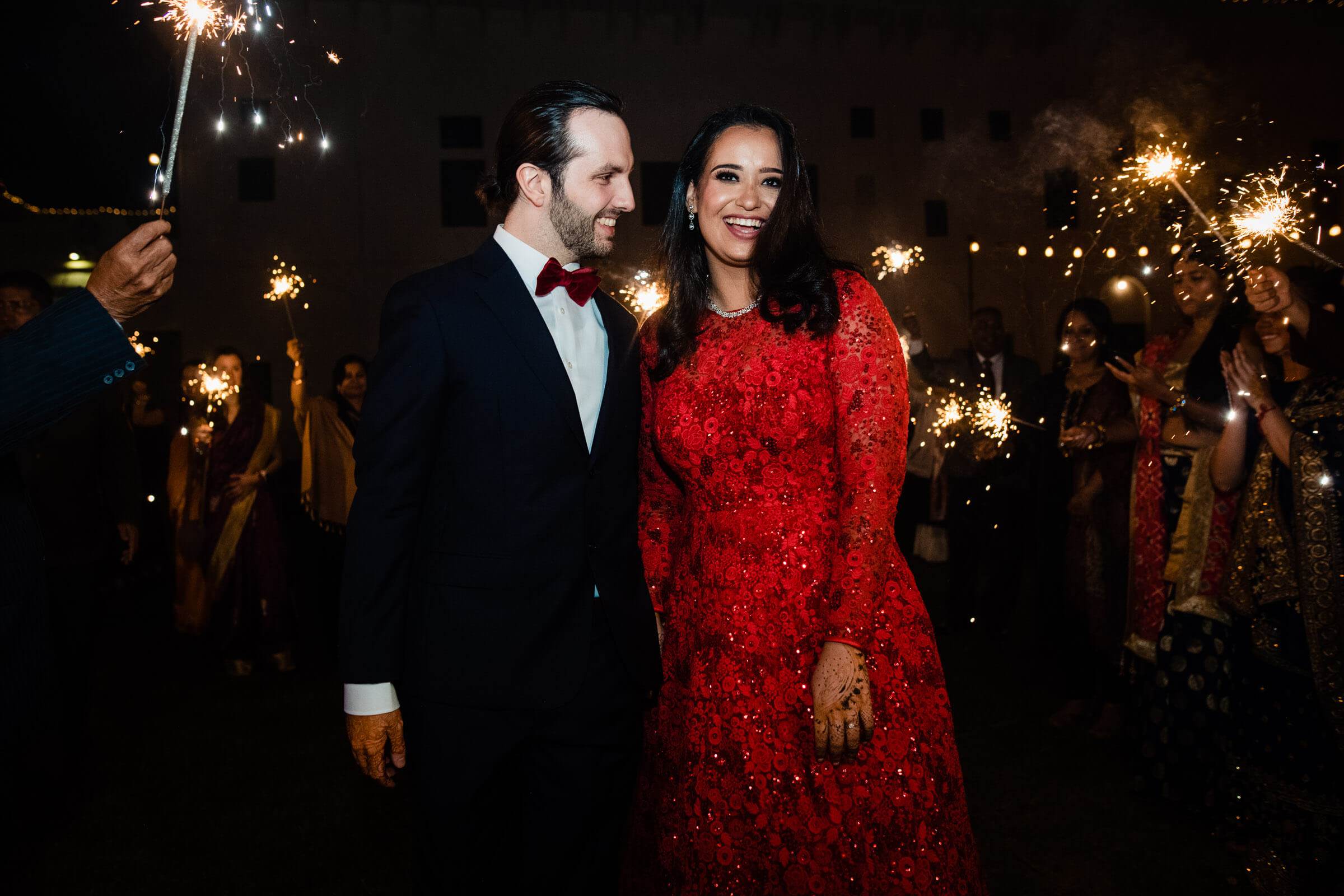 Maitrayee in red sequined gown and Eric in black tuxedo walk through sparkler-lined nighttime celebration in Rajasthan