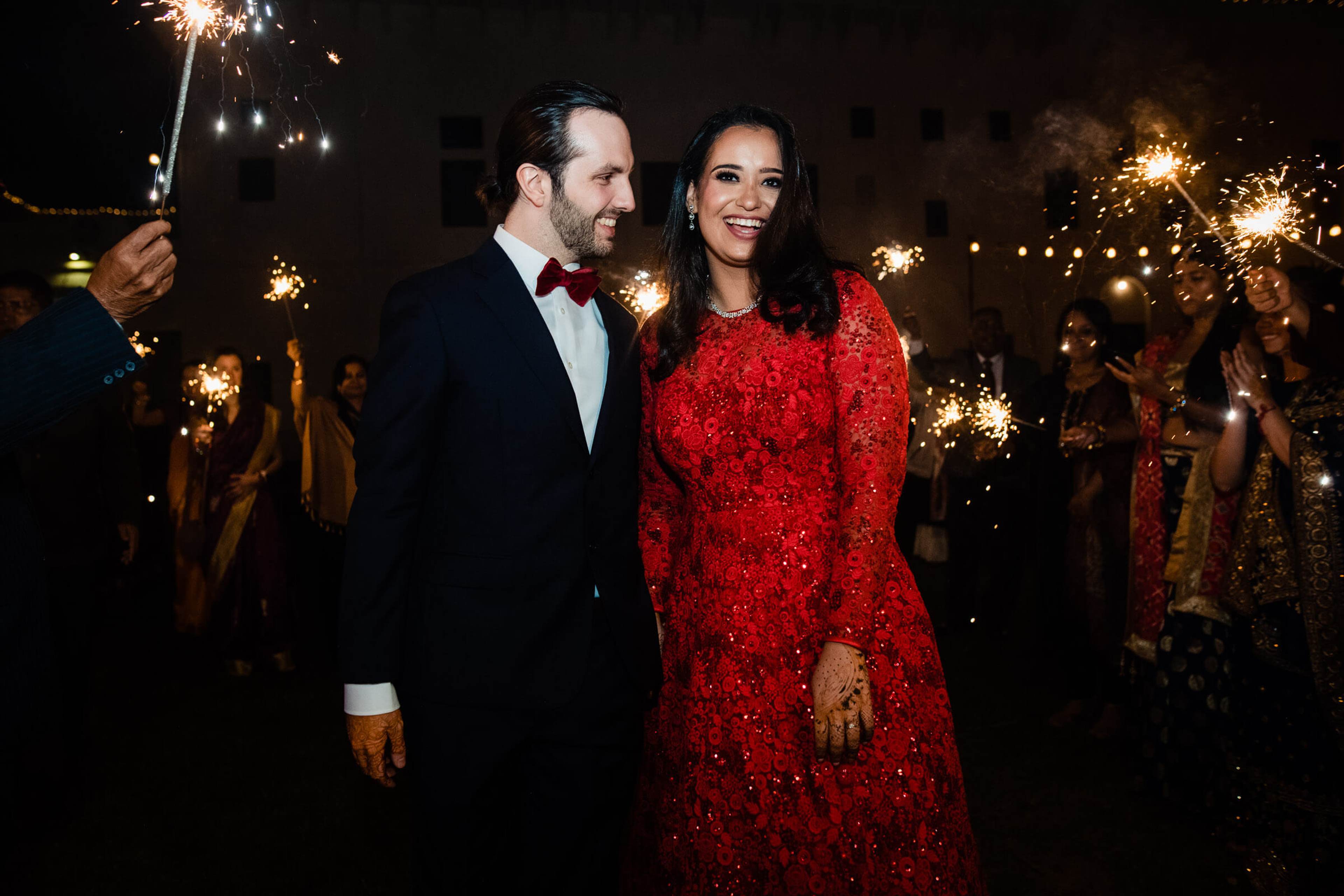 Maitrayee in red sequined gown and Eric in black tuxedo walk through sparkler-lined nighttime celebration in Rajasthan