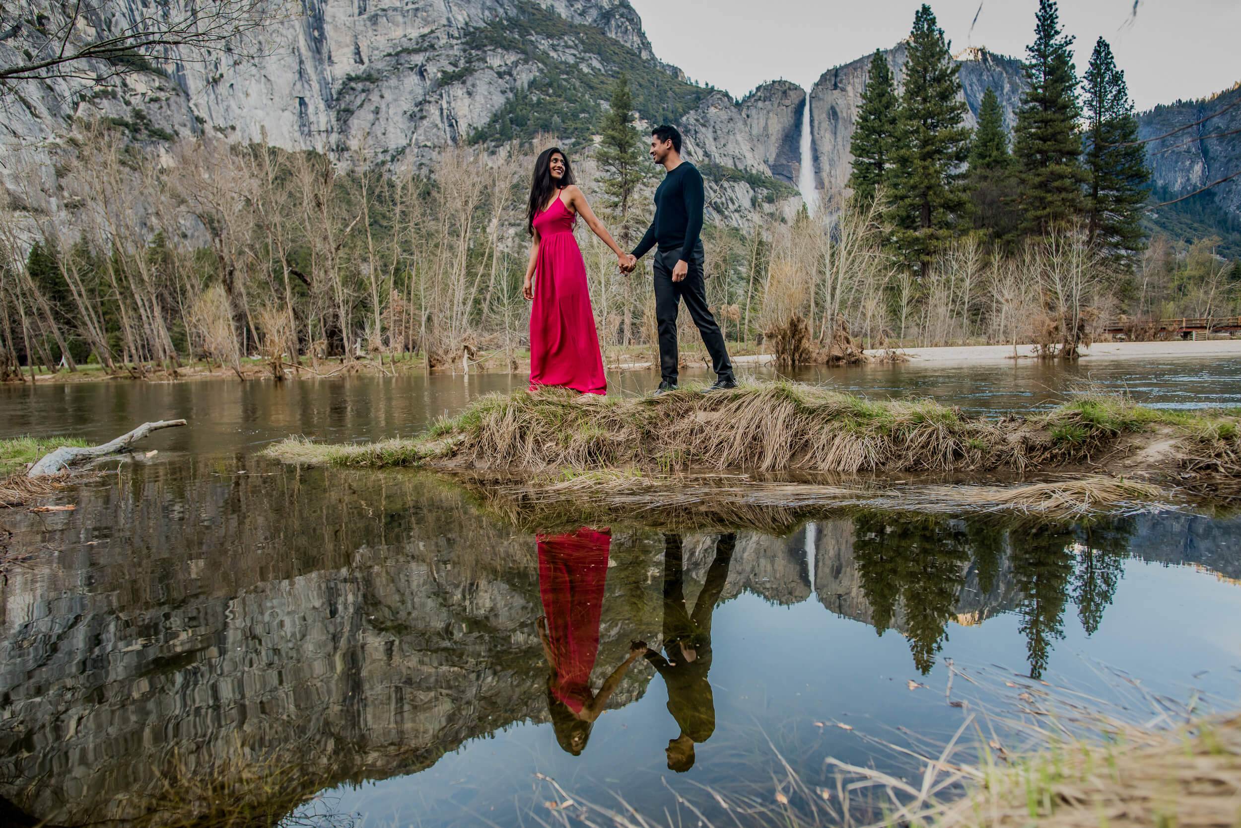 Anu and Raghu hold hands on a river bank, reflected in still water, with California cliffs and pine trees behind them