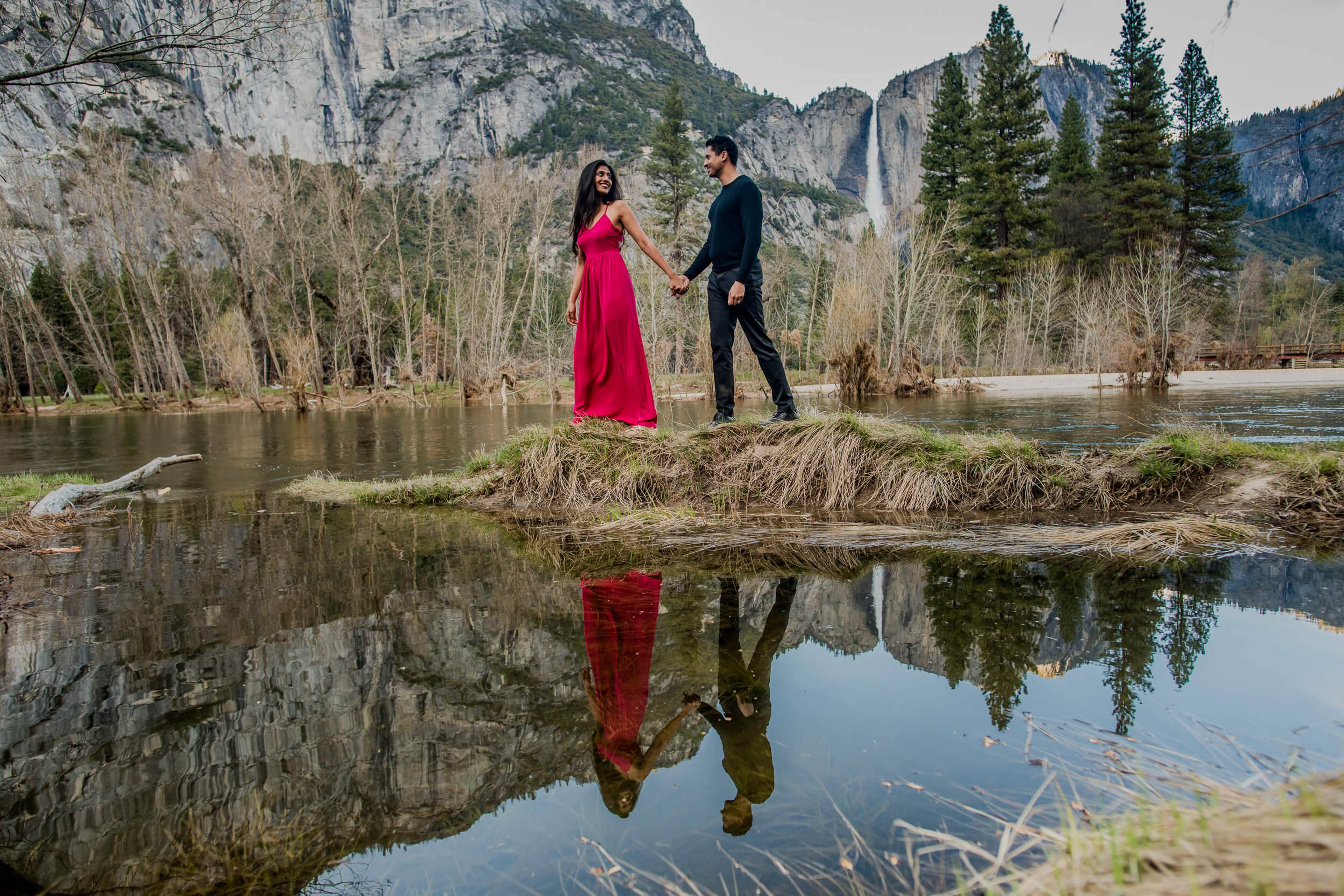 Anu and Raghu hold hands on a river bank, reflected in still water, with California cliffs and pine trees behind them