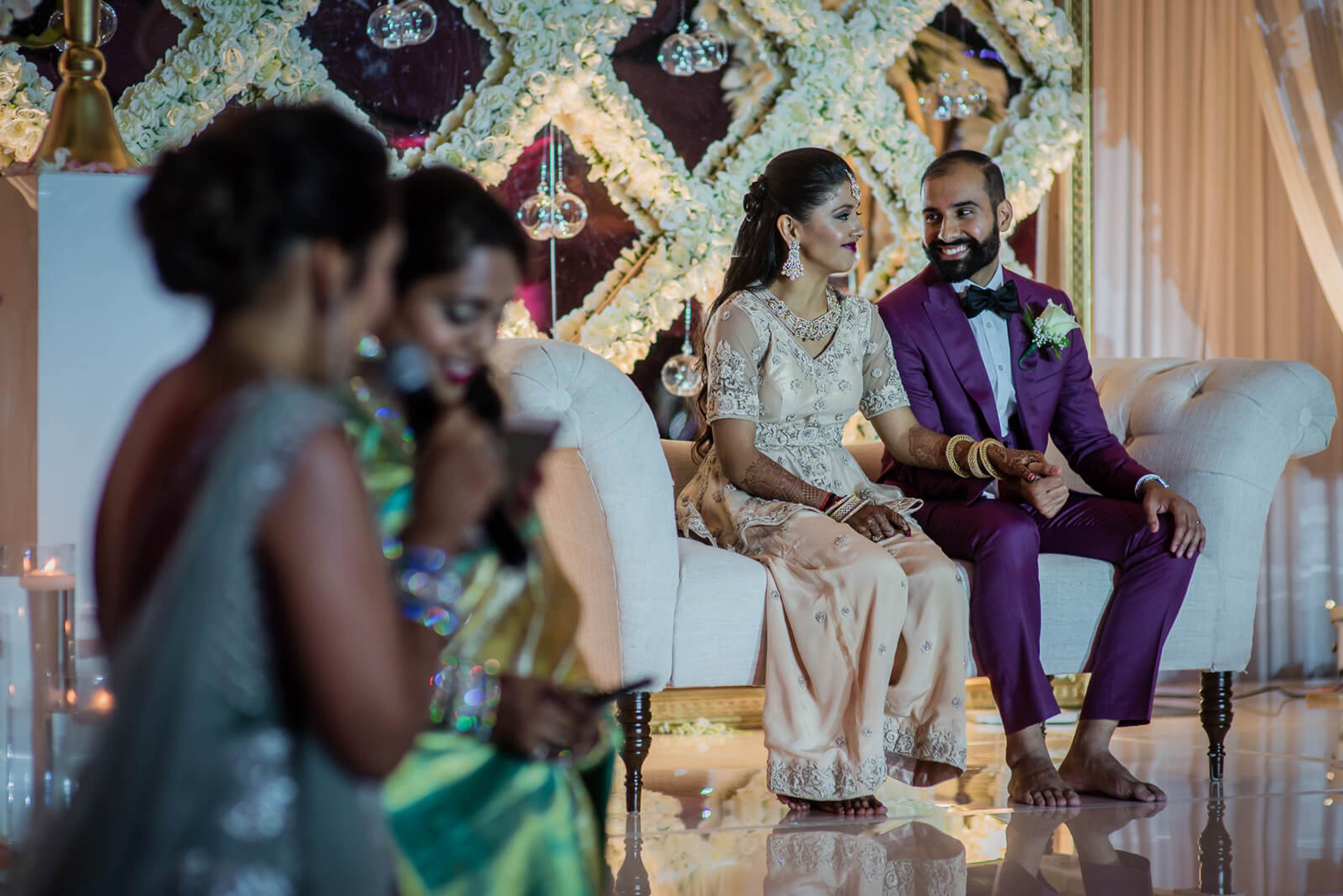Barkha and Ankit seated on white sofa in purple and gold wedding attire, ornate backdrop, guests in foreground