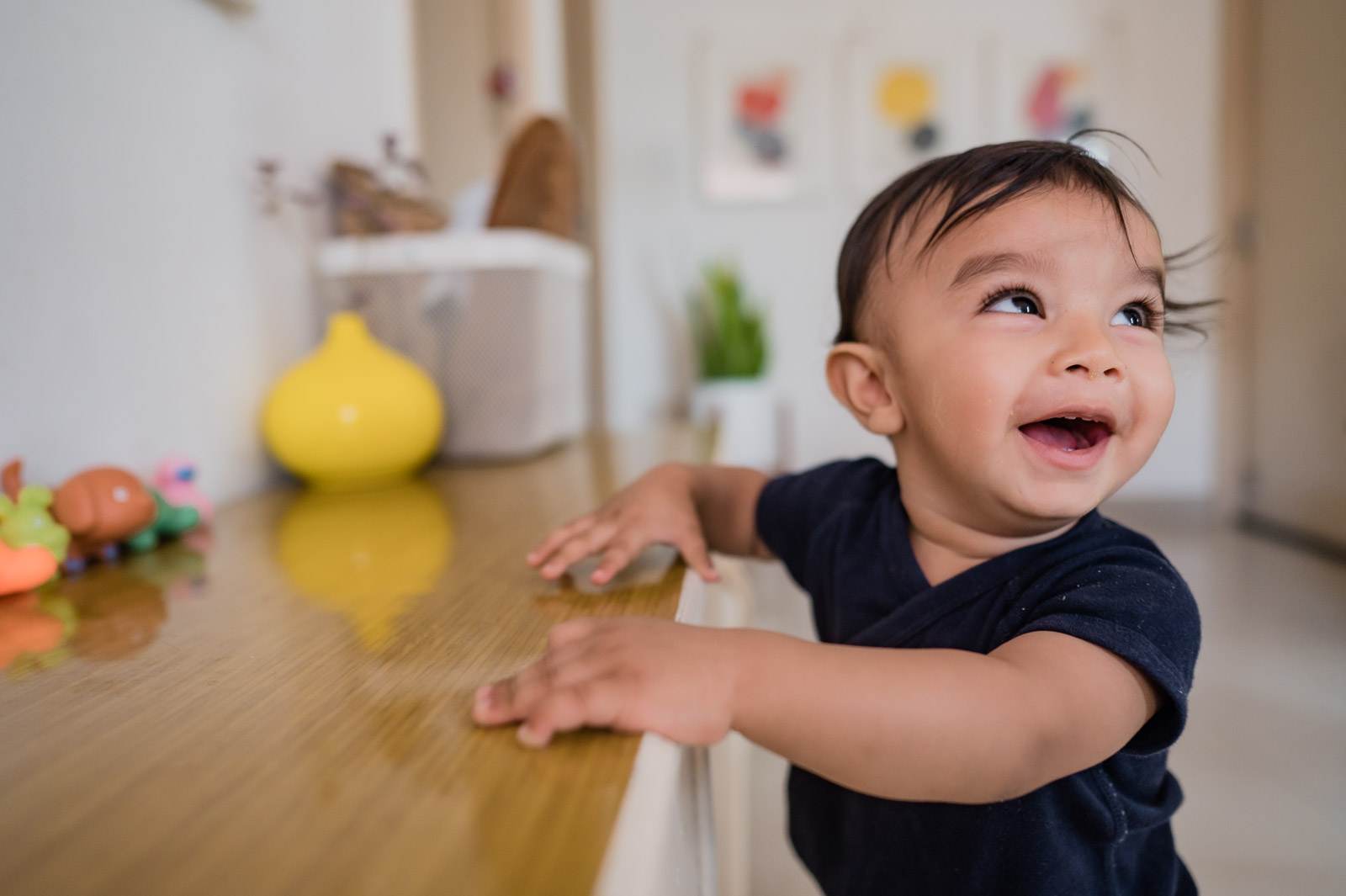 Toddler in black shirt smiling at table with toys and yellow bowl in bright kitchen