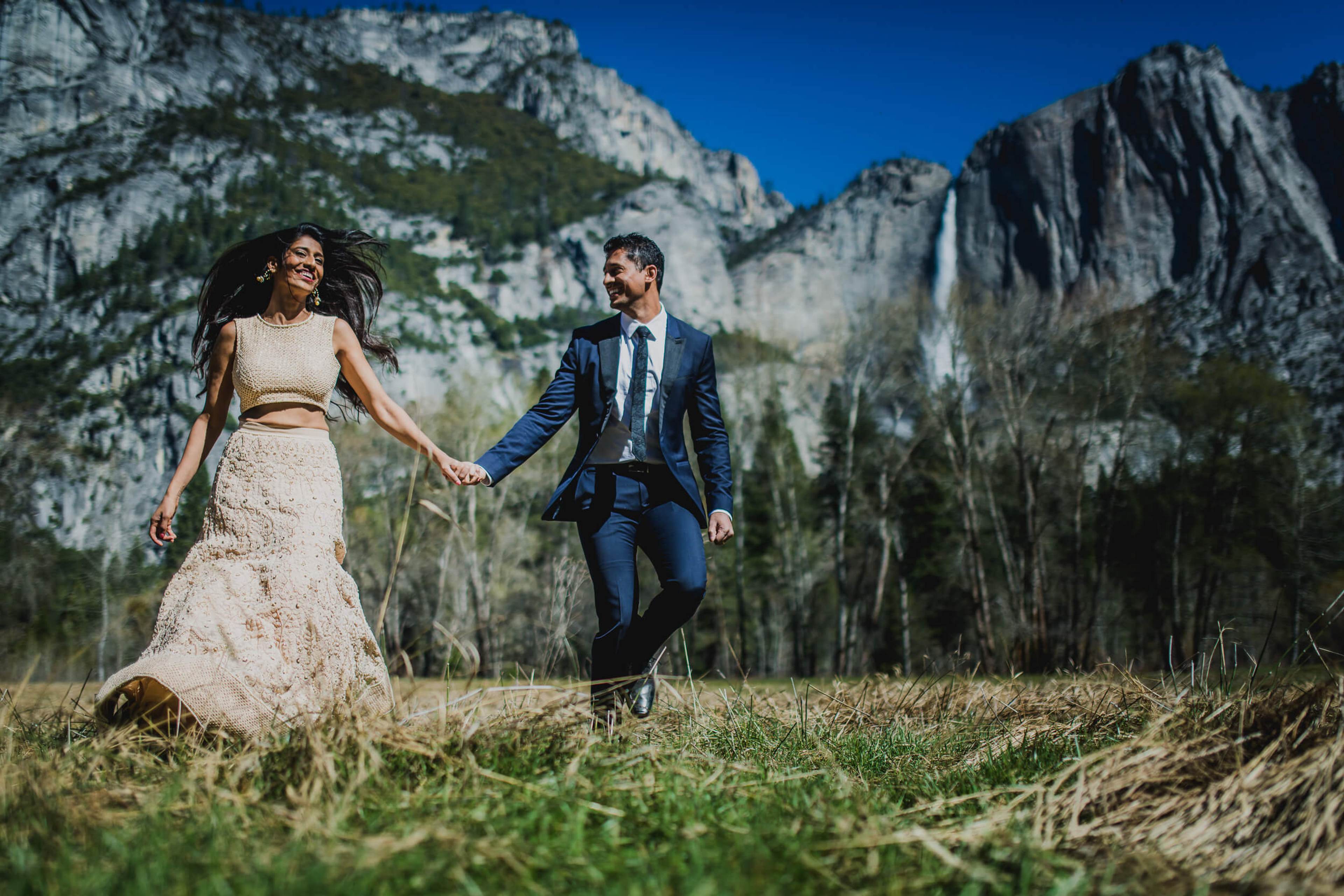 Anu and Raghu hold hands walking through a meadow with California's dramatic granite cliffs and waterfall behind them