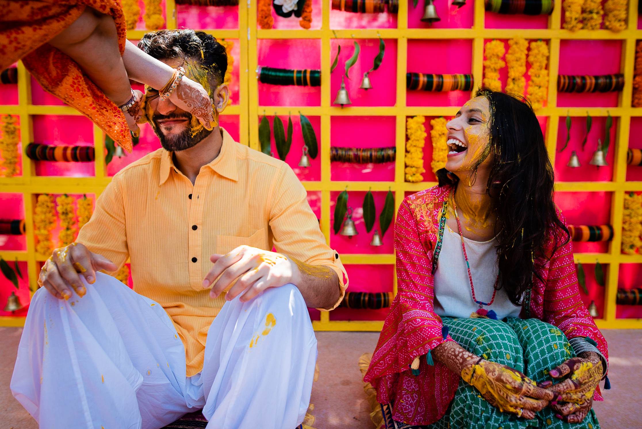 Avani and Srikant laugh joyfully against vibrant pink and yellow latticed backdrop in Goa