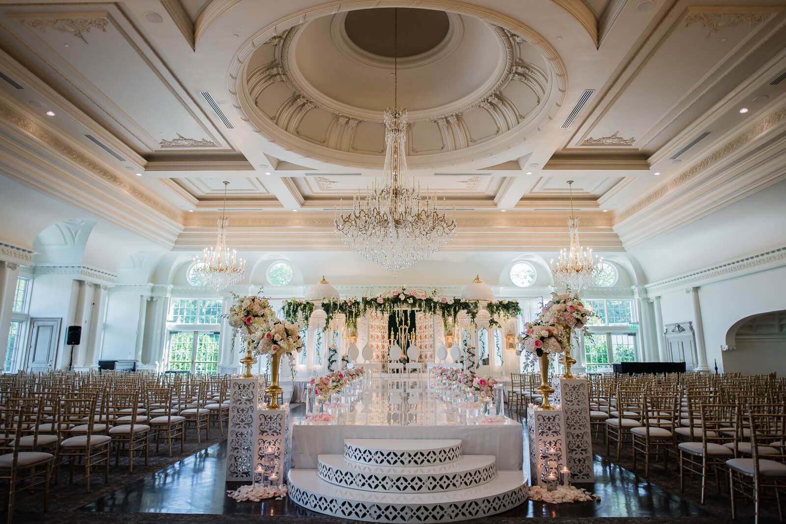 Elegant ballroom ceremony setup with ornate ceiling, crystal chandeliers, floral altar, and wooden chairs