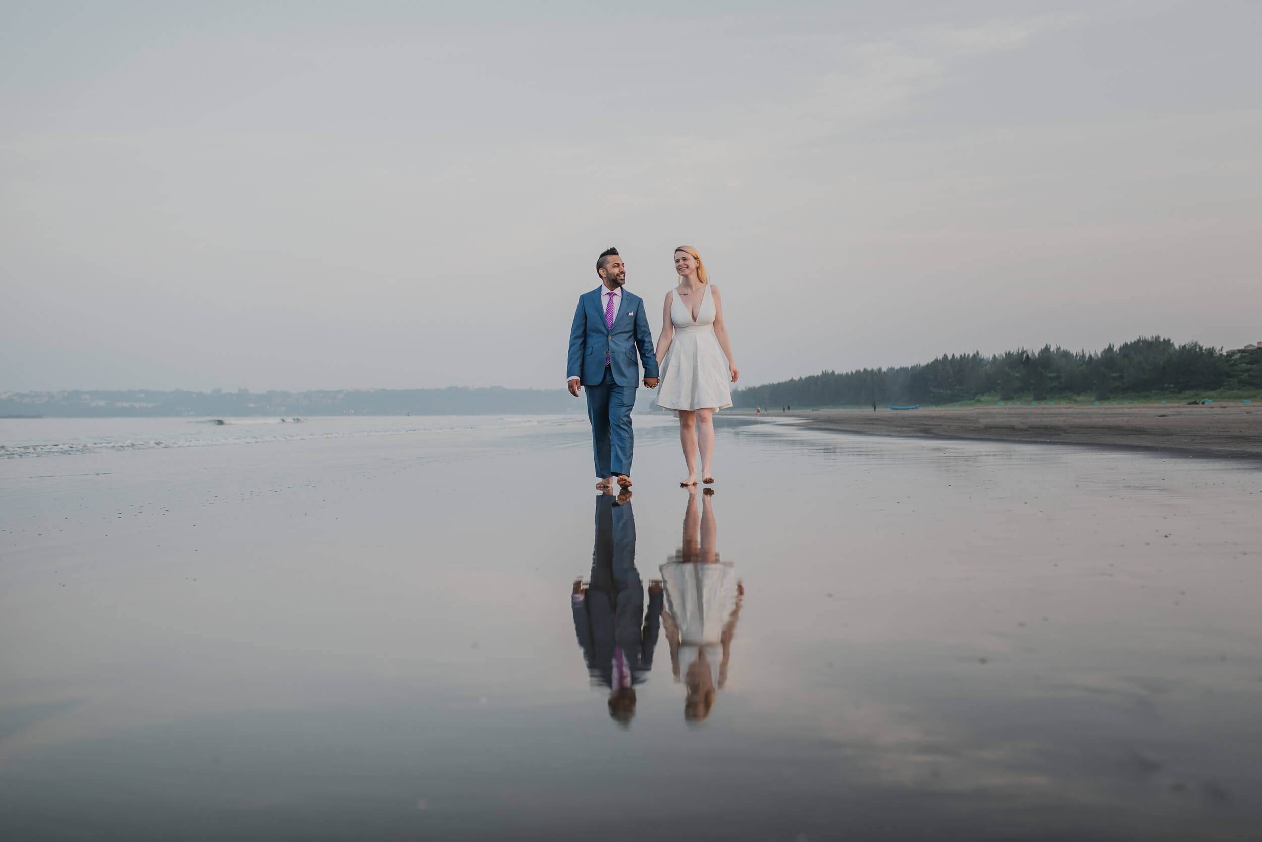 Sarah and Ravin walking hand in hand on reflective beach sand at Goa, mirrored in wet sand