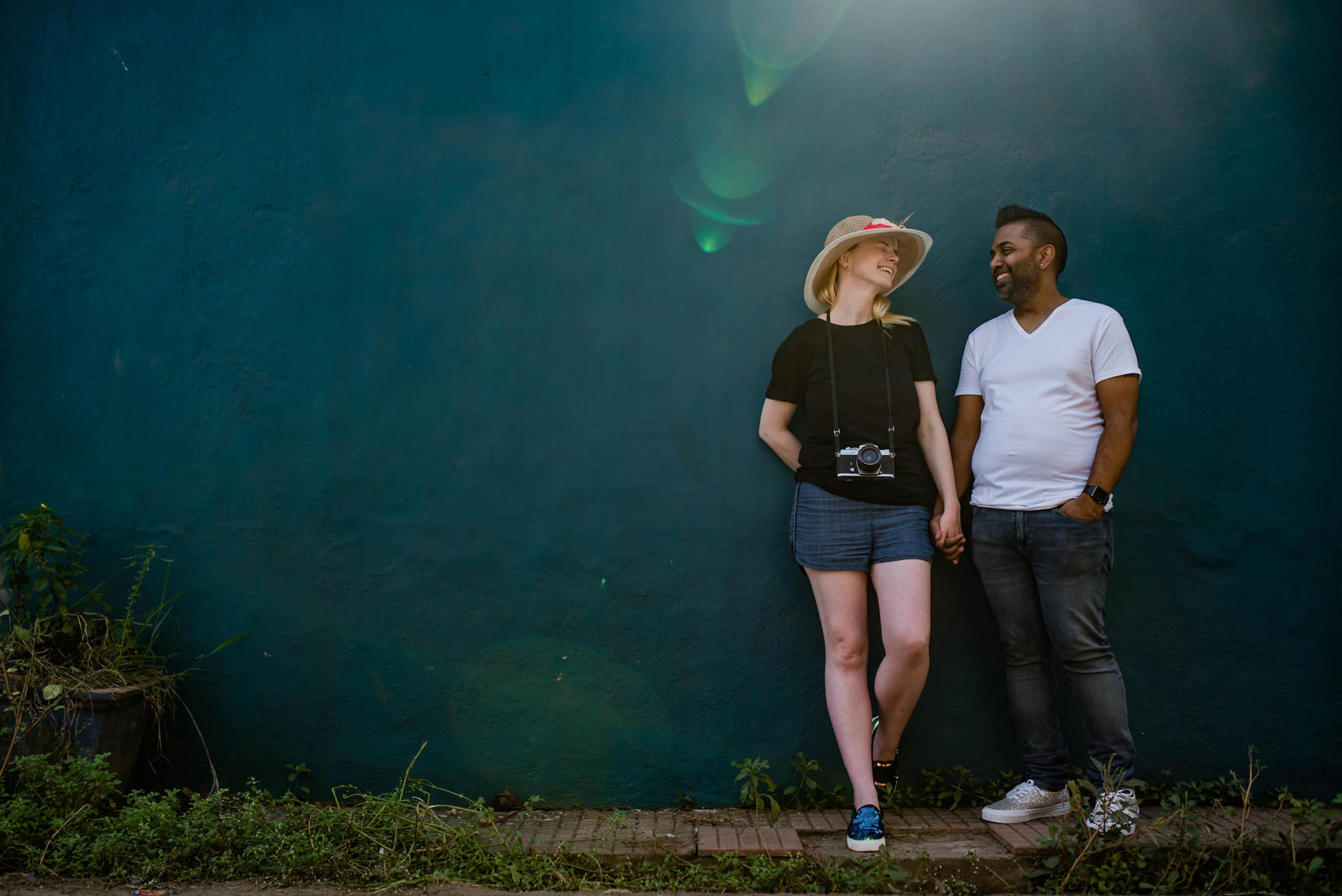Sarah and Ravin stand holding hands before a deep teal wall in Goa, backlit by soft green light