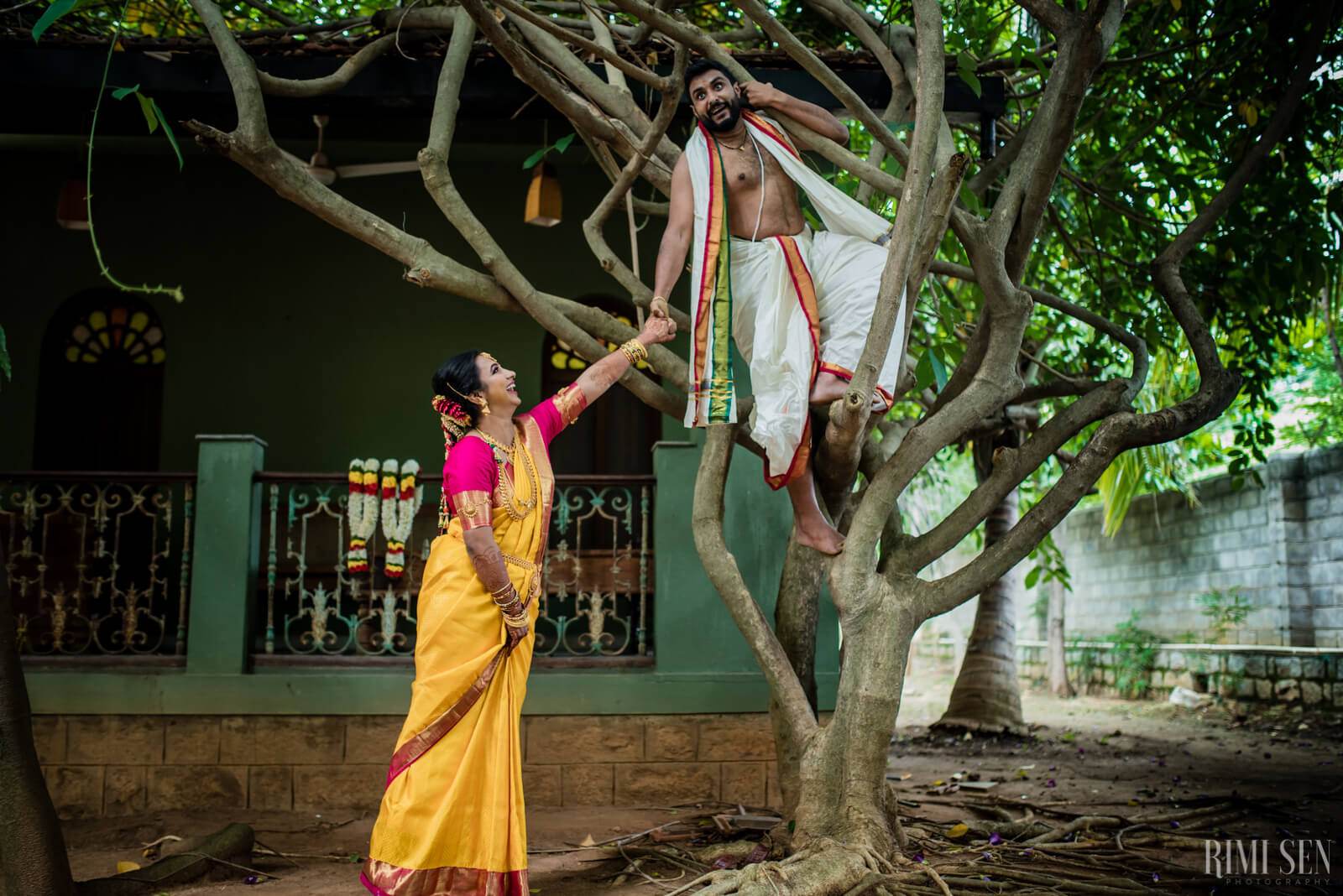 Nivetha in yellow saree reaching toward Sridhar seated in tree branches, traditional wedding portrait in Bangalore courtyard