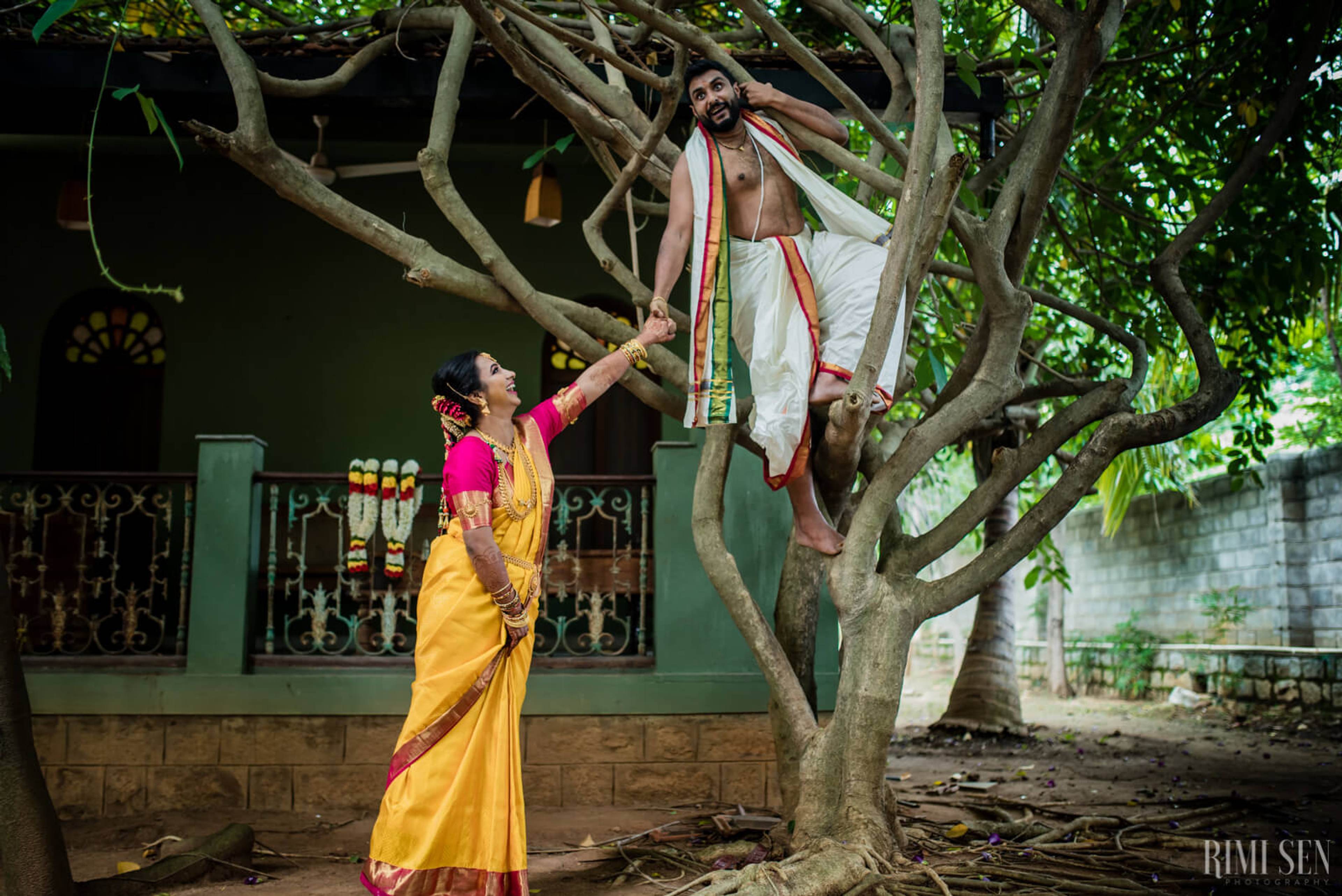 Nivetha in yellow saree reaching toward Sridhar seated in tree branches, traditional wedding portrait in Bangalore courtyard