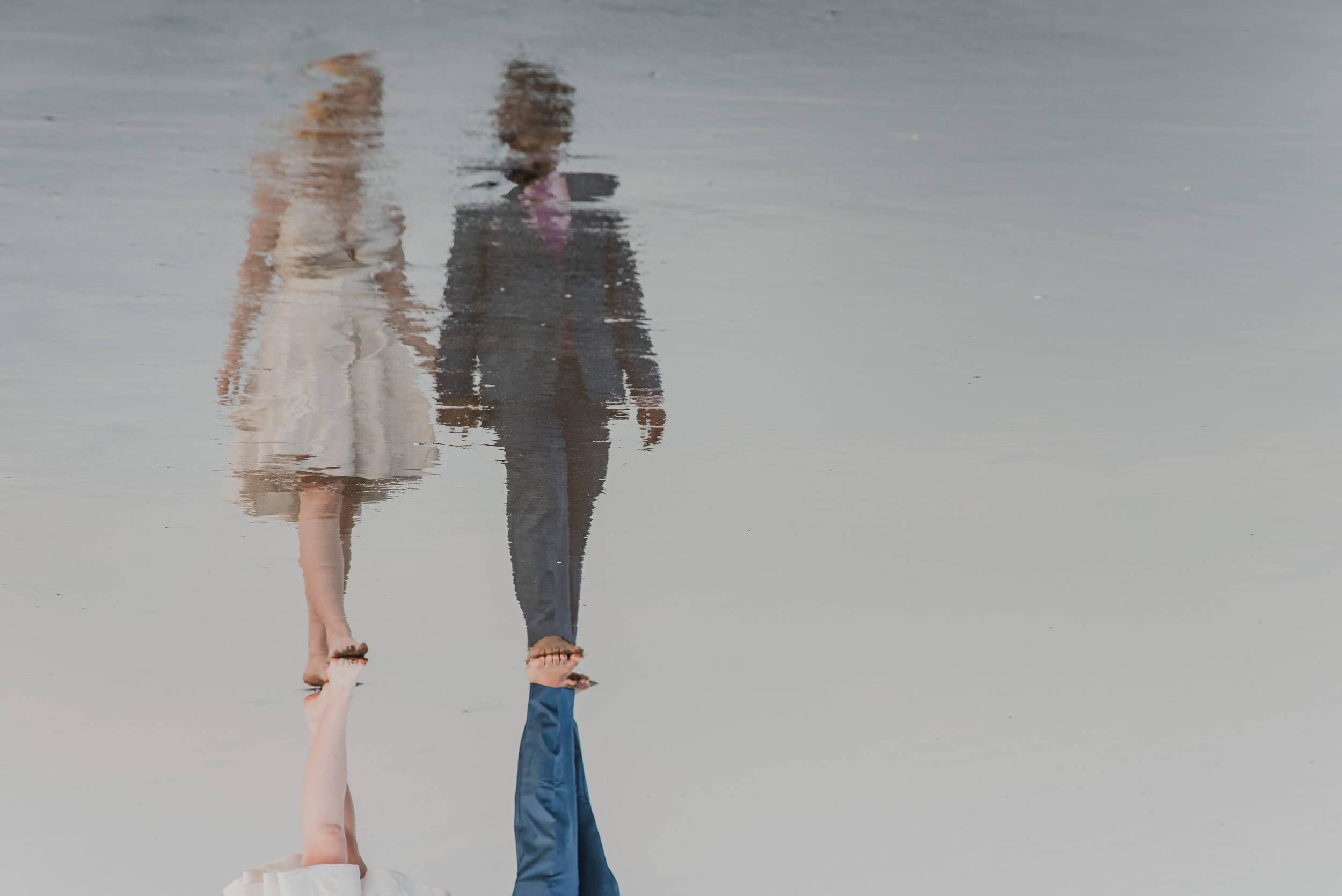 Couple holding hands reflected in water, woman in cream dress, man in dark suit, Goa