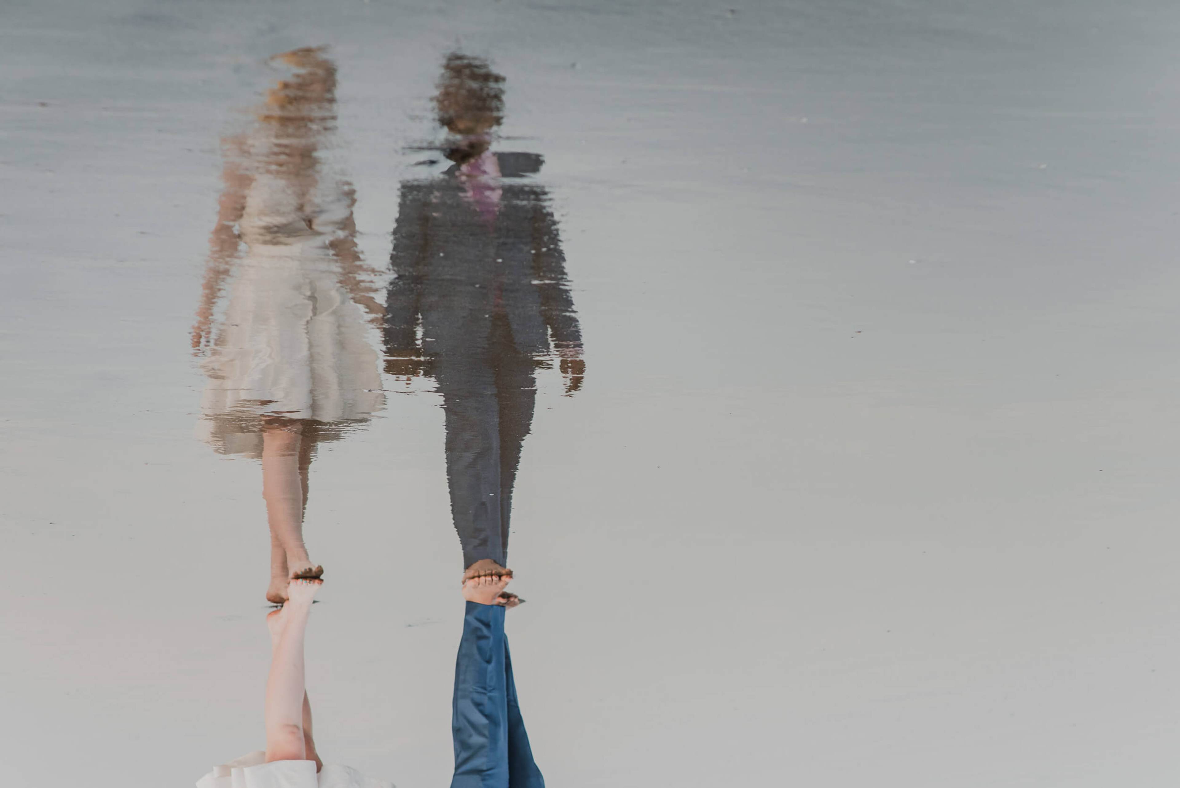 Couple holding hands reflected in water, woman in cream dress, man in dark suit, Goa