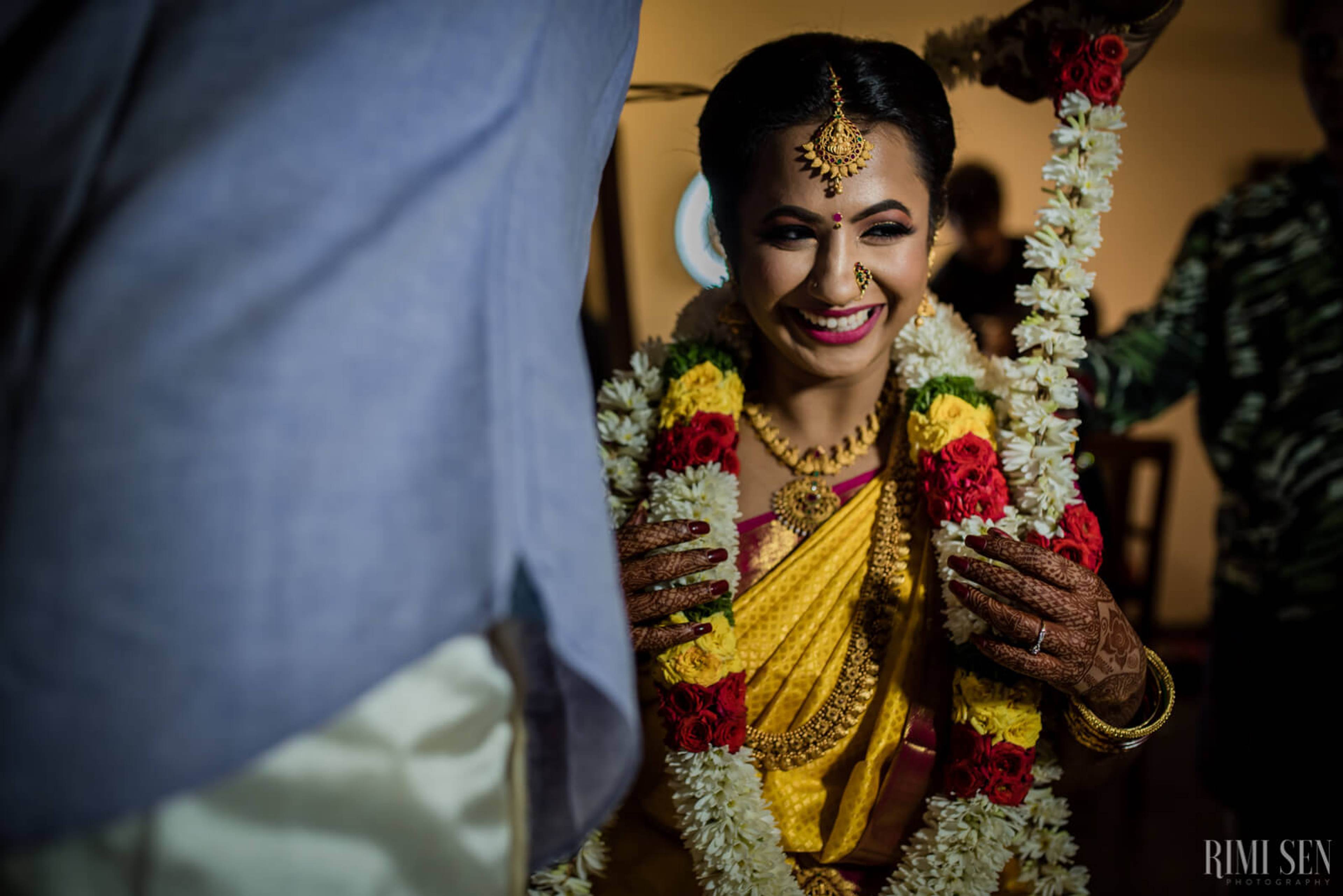 Nivetha smiles in traditional yellow silk and ornate gold jewelry, draped in flower garlands at Bangalore wedding