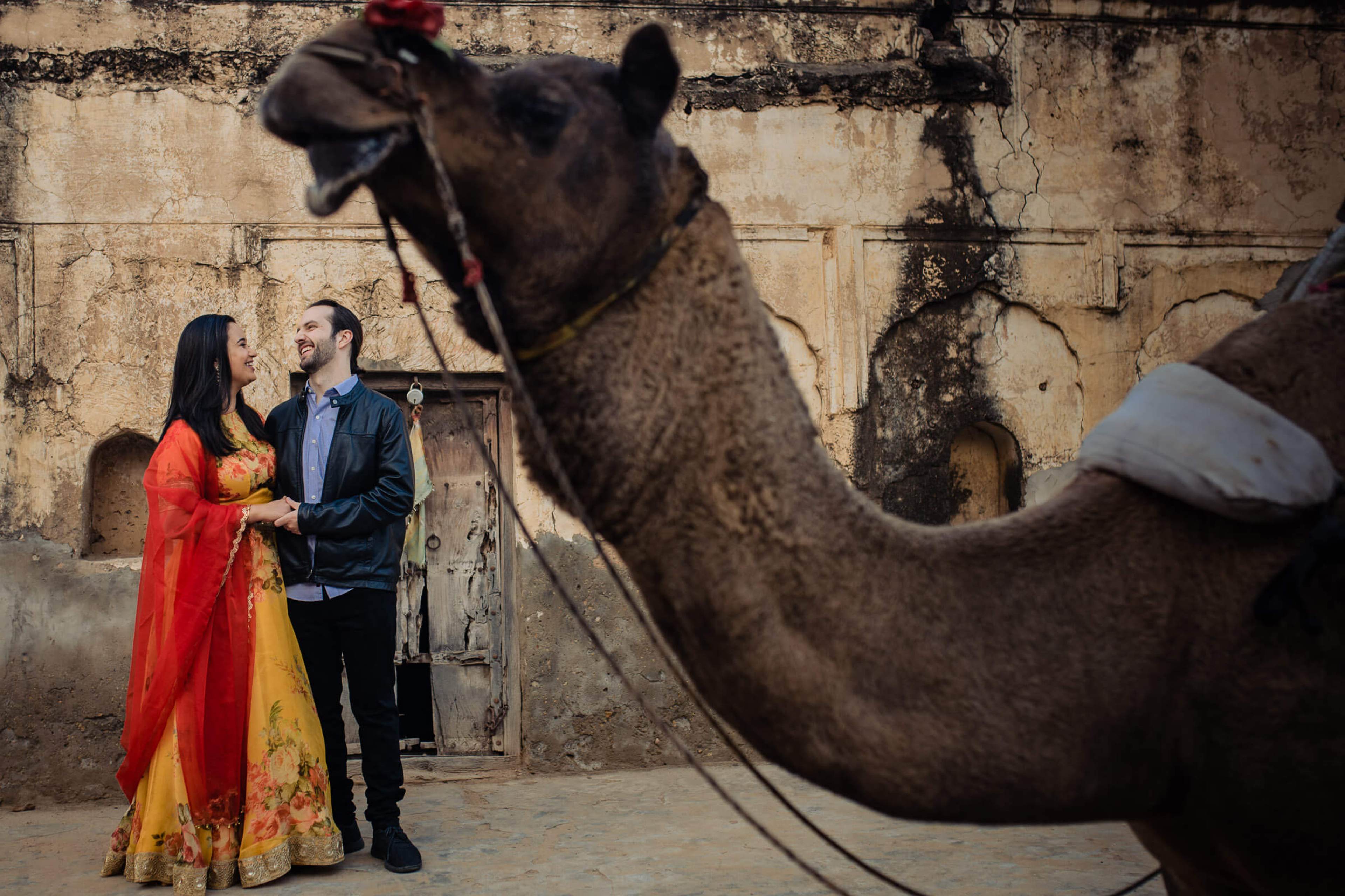 Maitrayee and Eric stand together beside a camel against weathered stone walls in Rajasthan