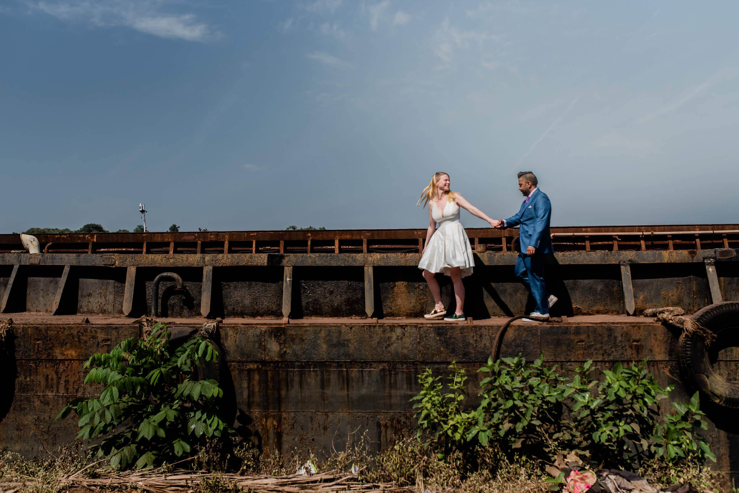 Sarah and Ravin holding hands on weathered fortification wall in Goa, blue sky behind