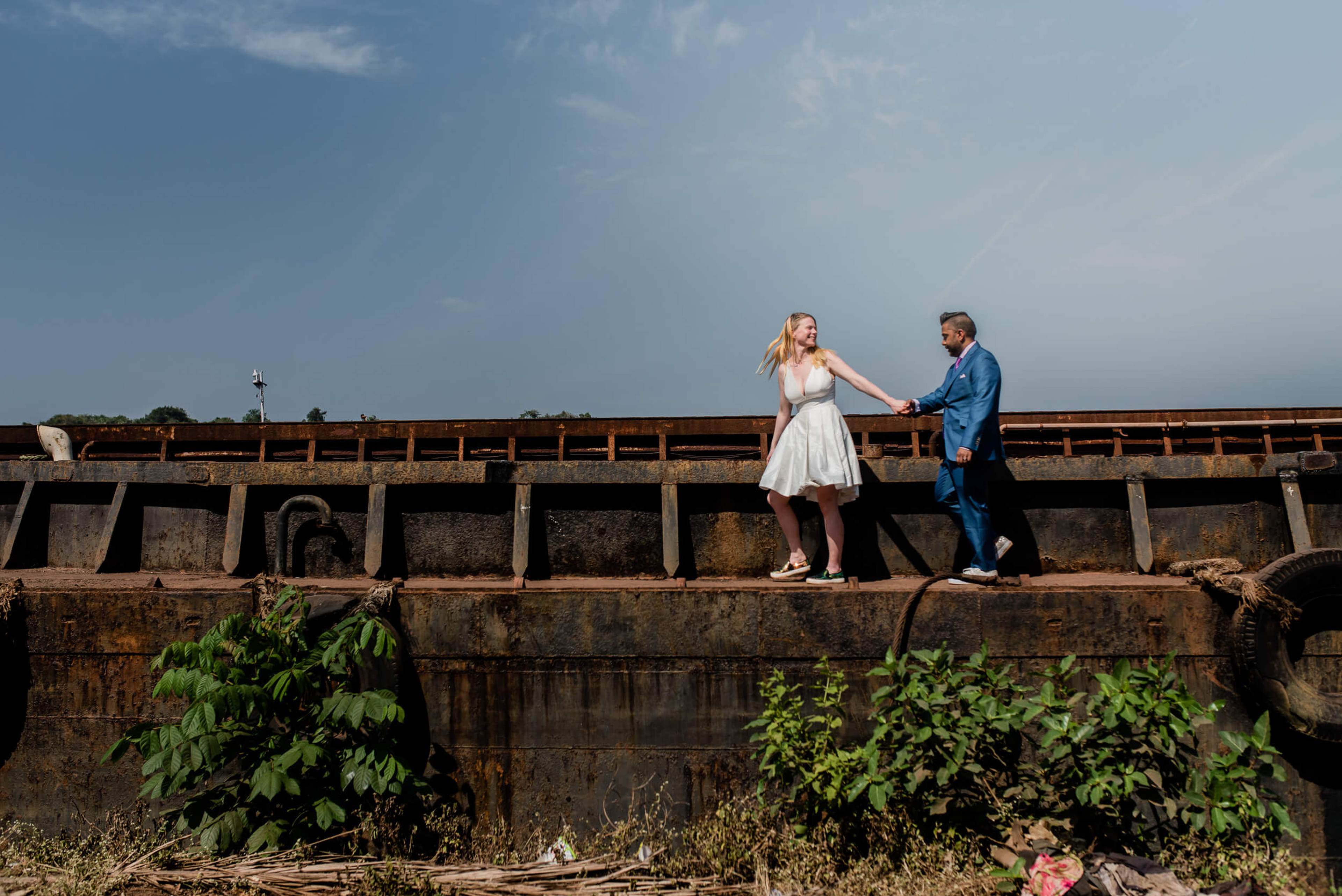 Sarah and Ravin holding hands on weathered fortification wall in Goa, blue sky behind