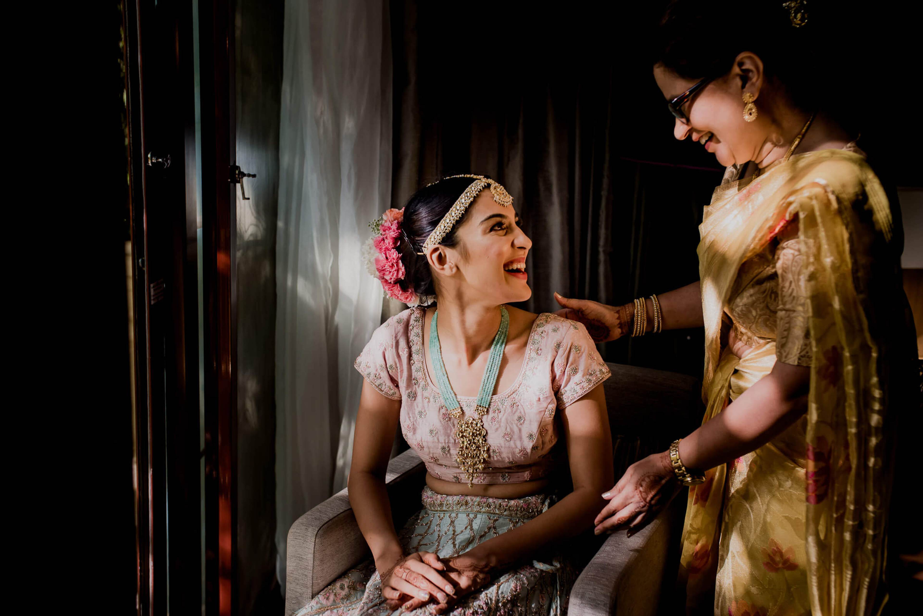 Woman in ornate white blouse with pink flowers in hair sits while another woman in gold sari adjusts her clothing indoors