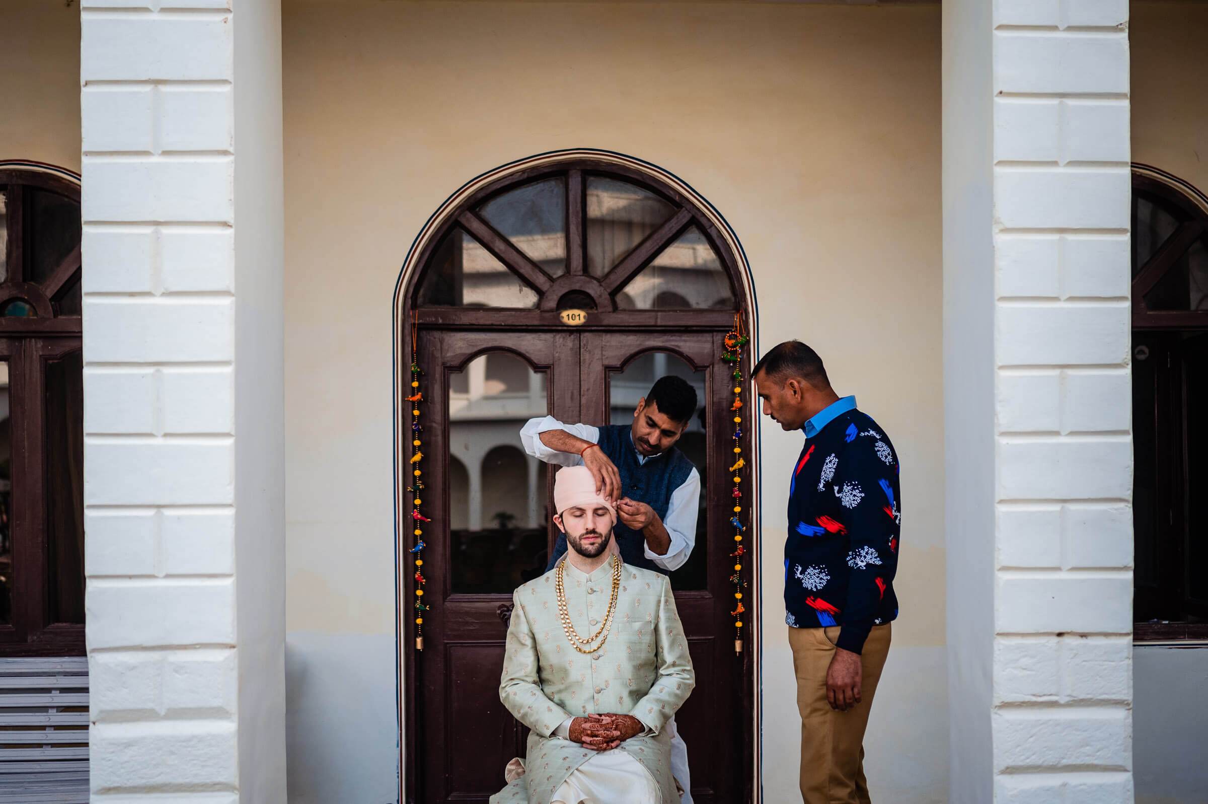 Maitrayee sits in traditional attire at an ornate arched doorway in Rajasthan while two men adjust his appearance