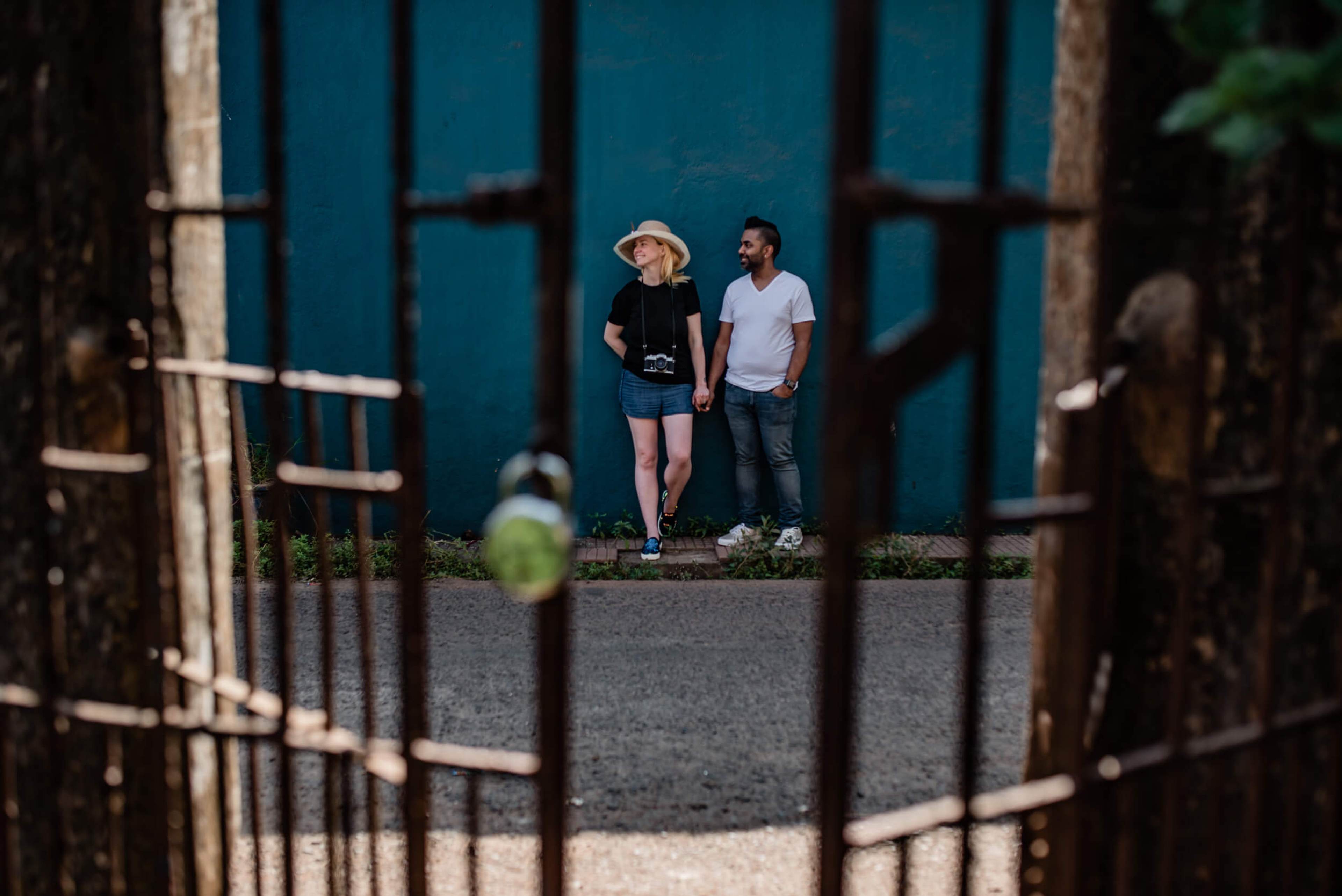 Sarah and Ravin walk together against a teal building, framed through a rusty gate in Goa