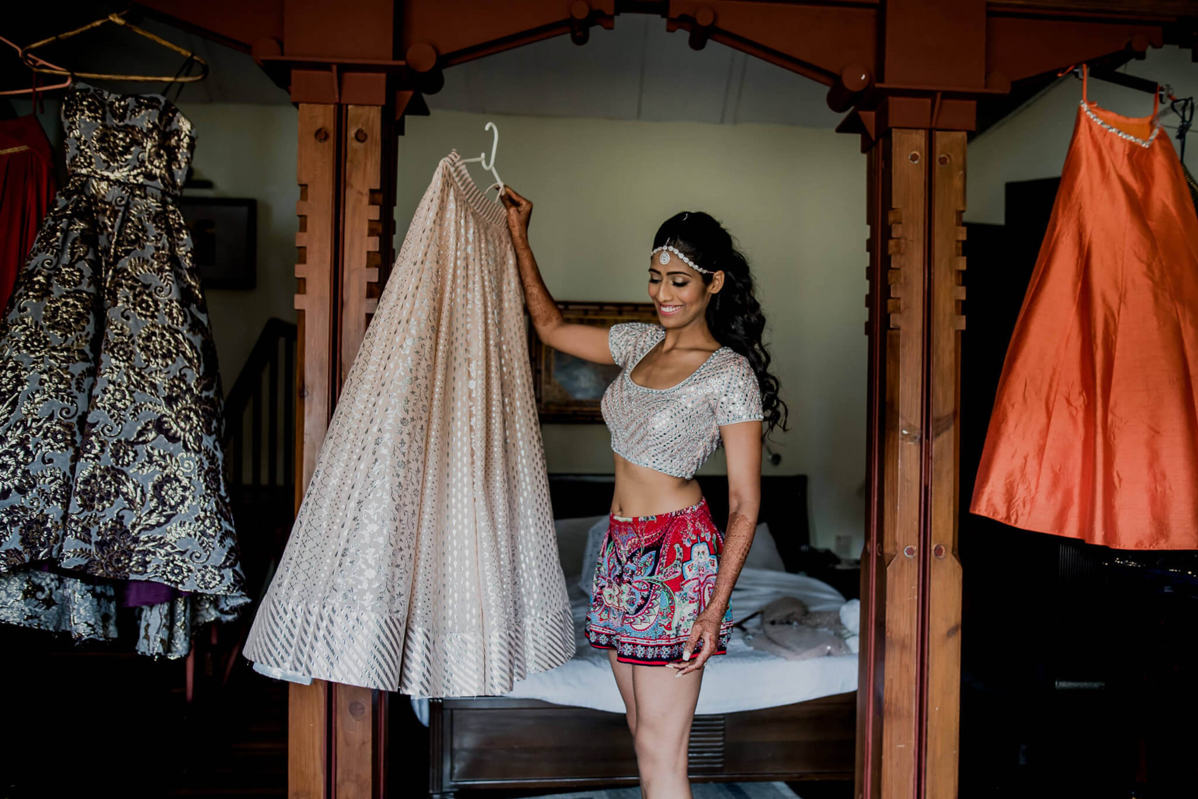 Anu holding delicate veil in traditional bedroom with ornate wooden frame and colorful garments hanging