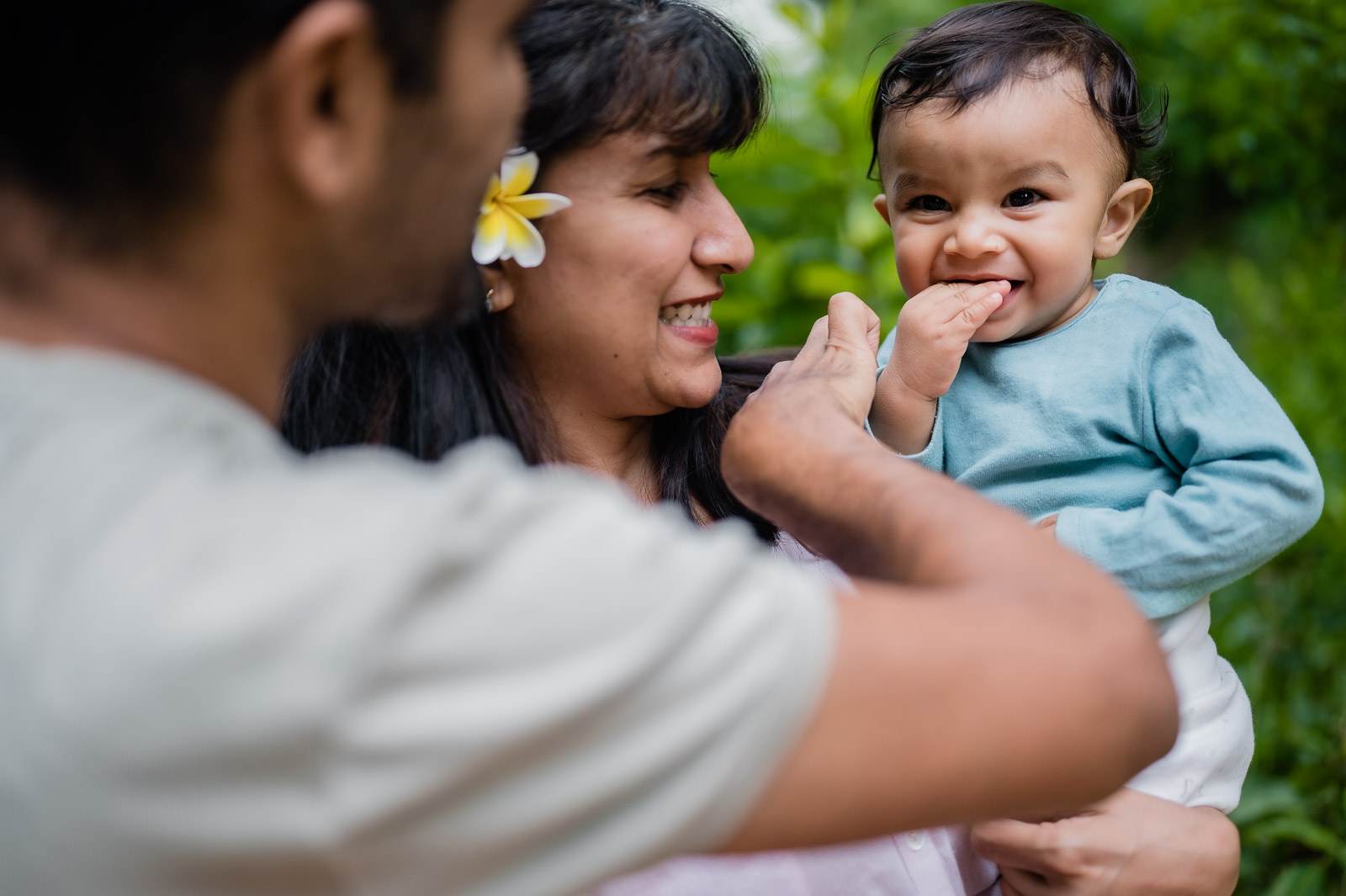 Woman with yellow flower in hair smiles while holding toddler, man in white shirt beside them outdoors