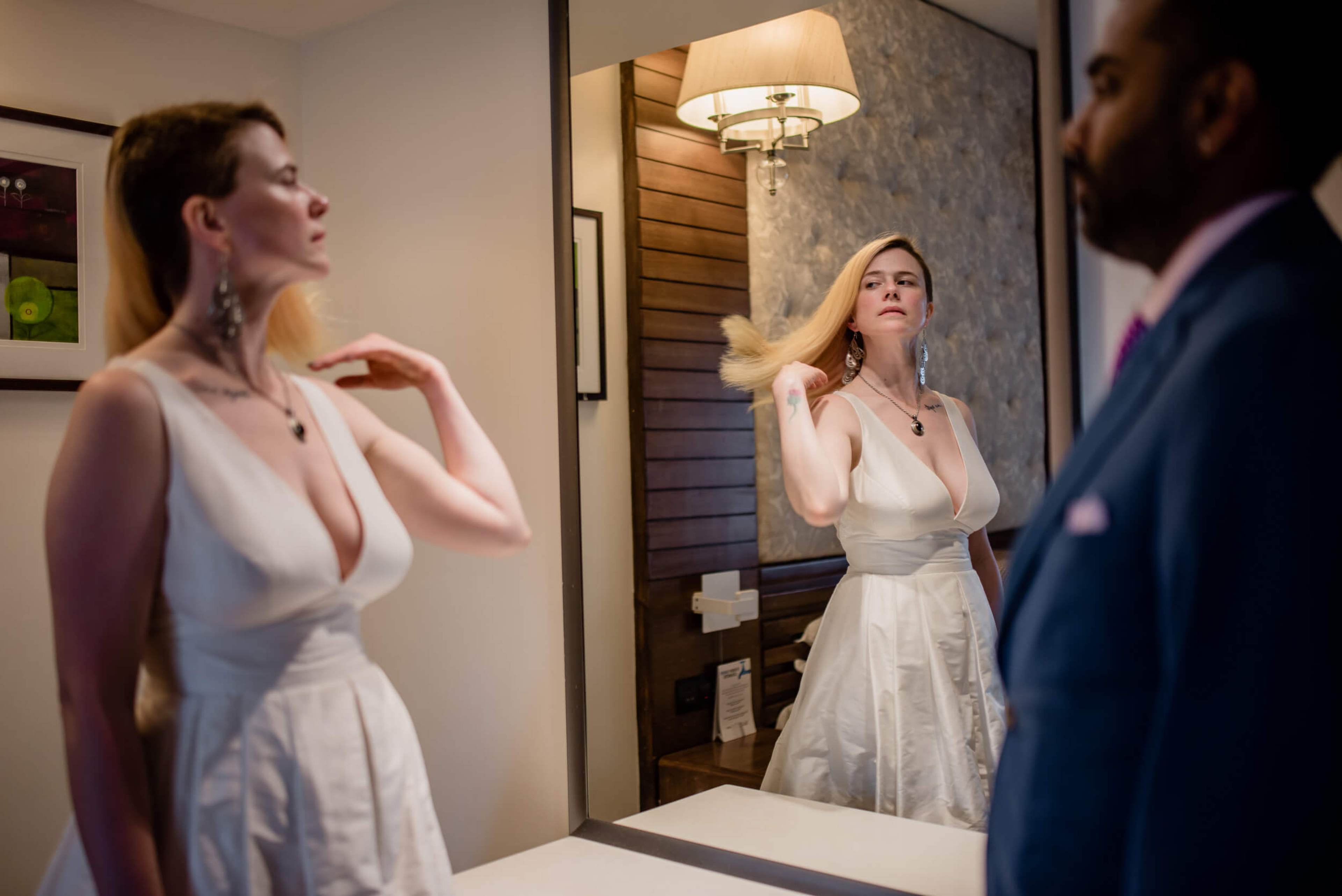 Sarah in white dress adjusting jewelry before mirror, Ravin partially visible in foreground, warm indoor lighting