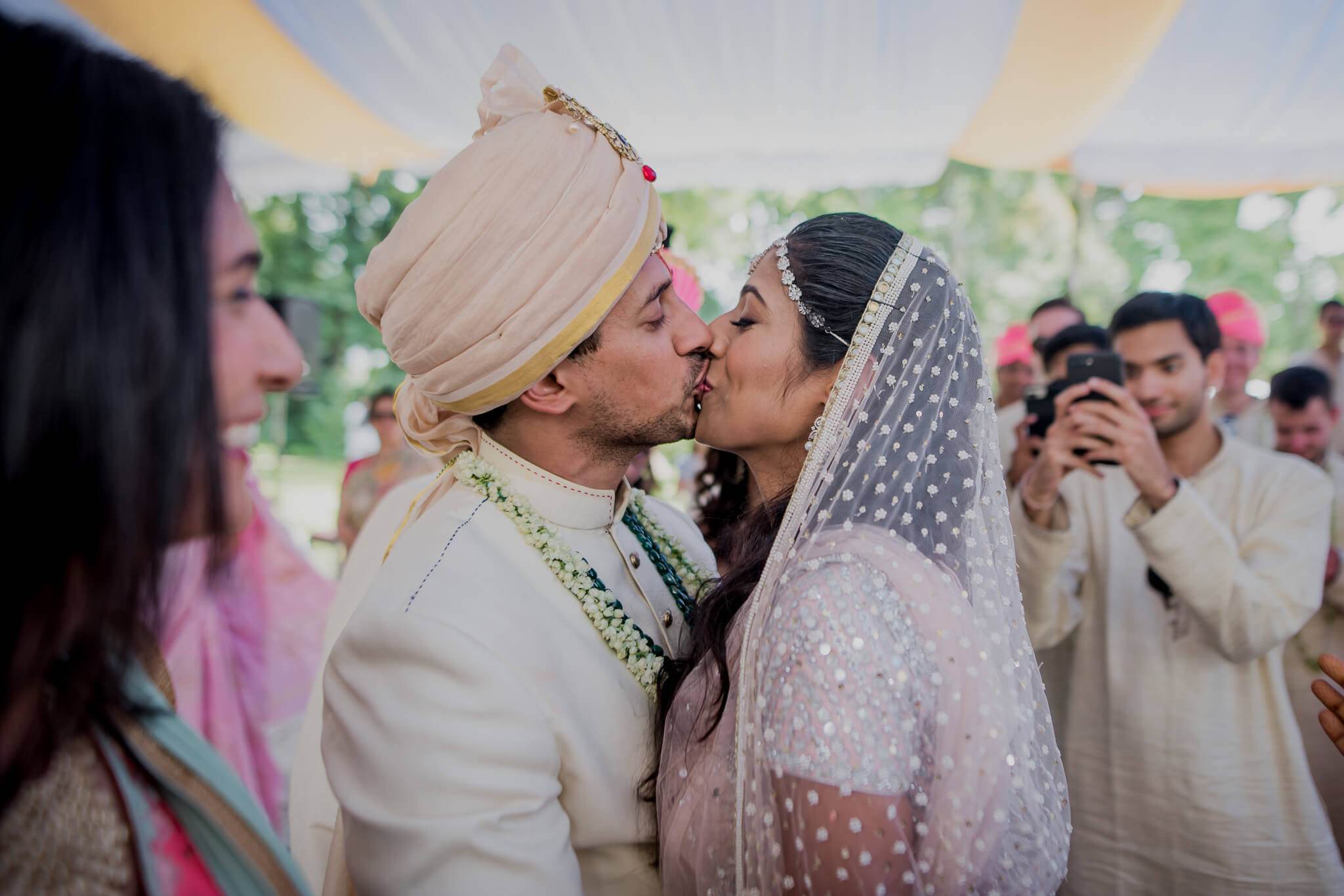 Anu and Raghu kiss under a tent during their wedding ceremony in Nepal, surrounded by guests in traditional attire