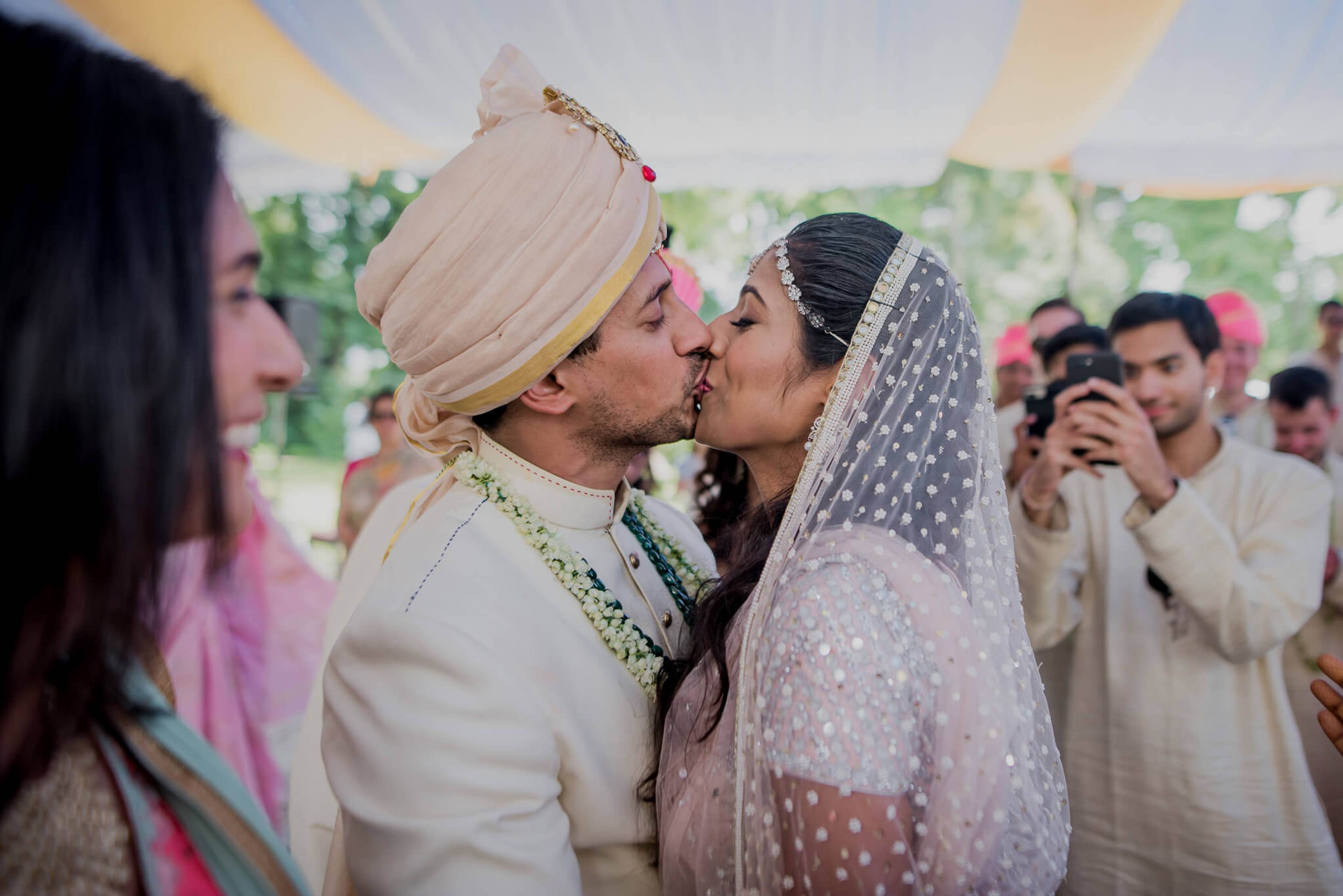 Anu and Raghu kiss under a tent during their wedding ceremony in Nepal, surrounded by guests in traditional attire