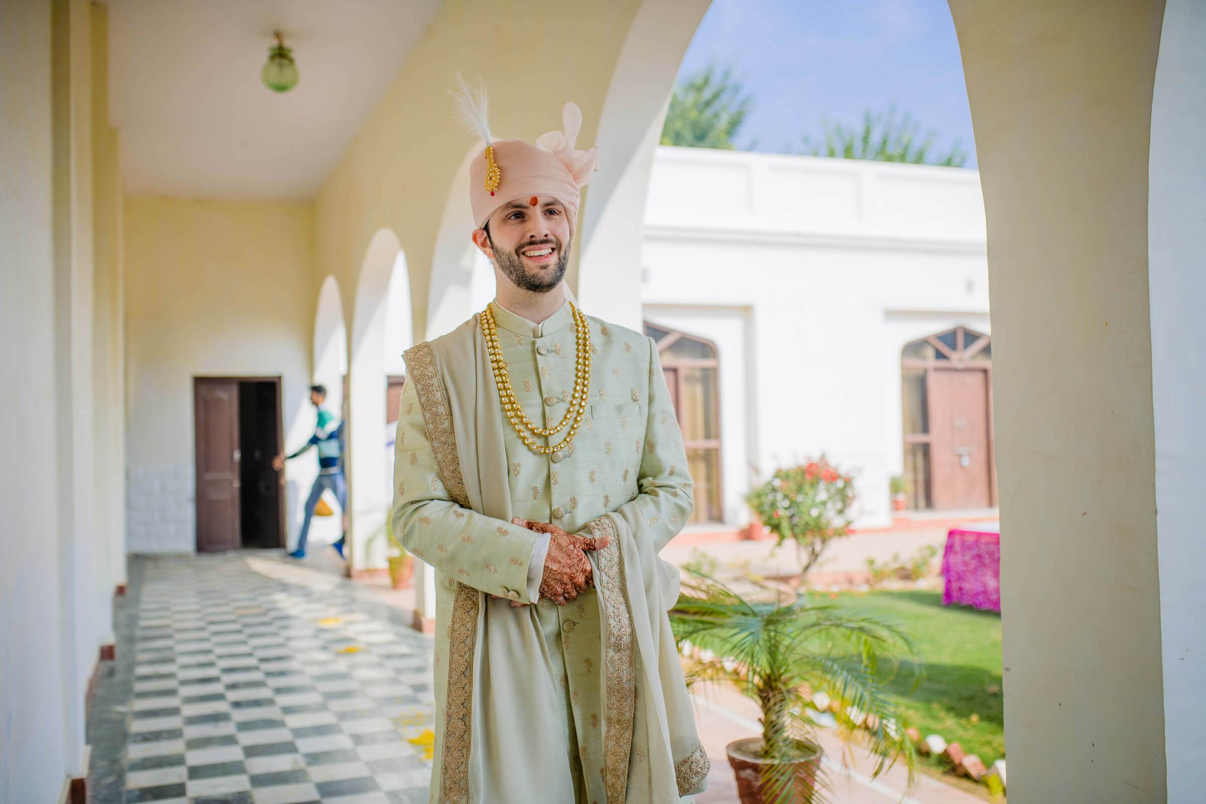 Groom Eric in traditional cream sherwani and turban poses in arched Rajasthan corridor with courtyard beyond