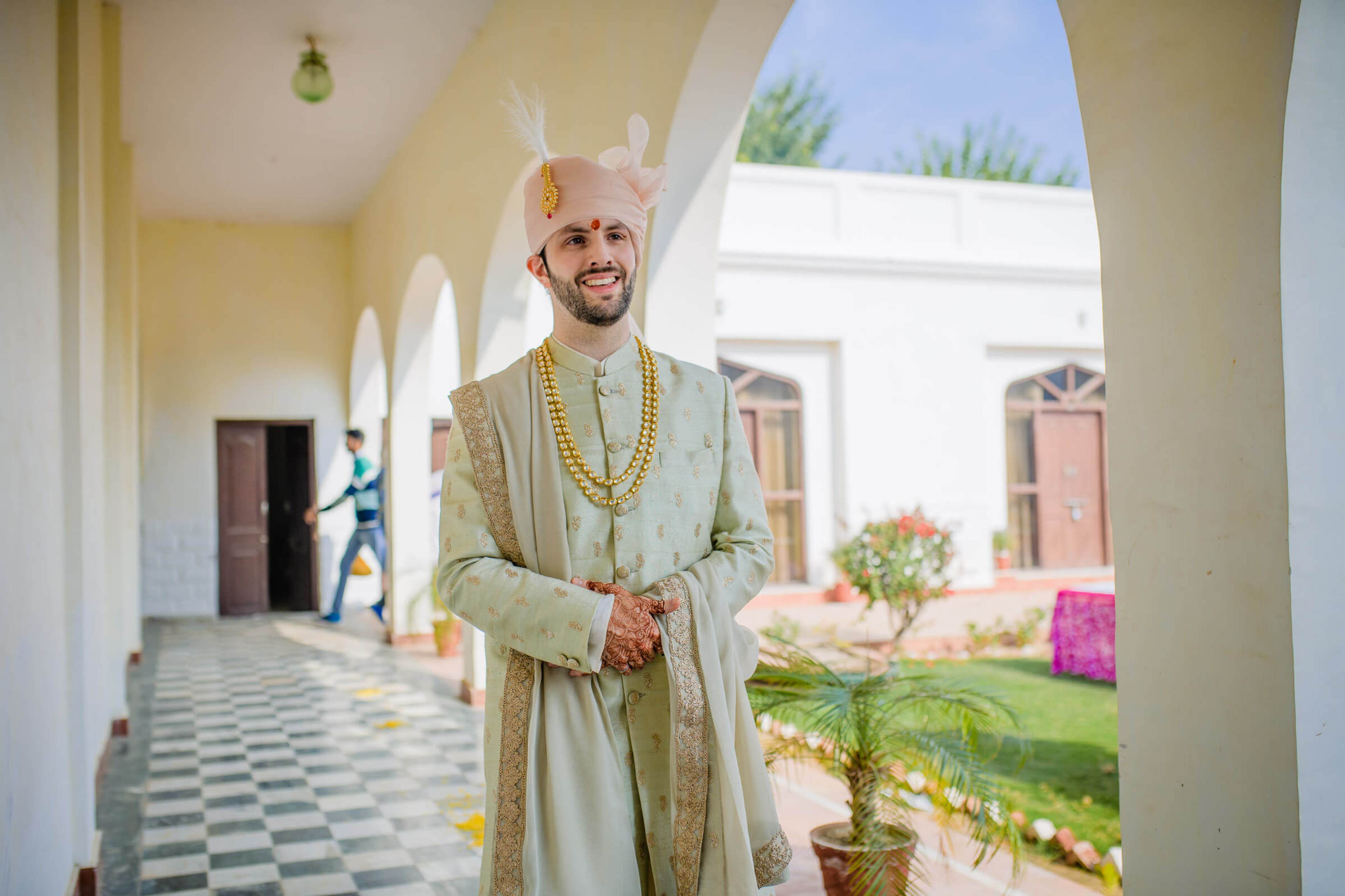 Groom Eric in traditional cream sherwani and turban poses in arched Rajasthan corridor with courtyard beyond